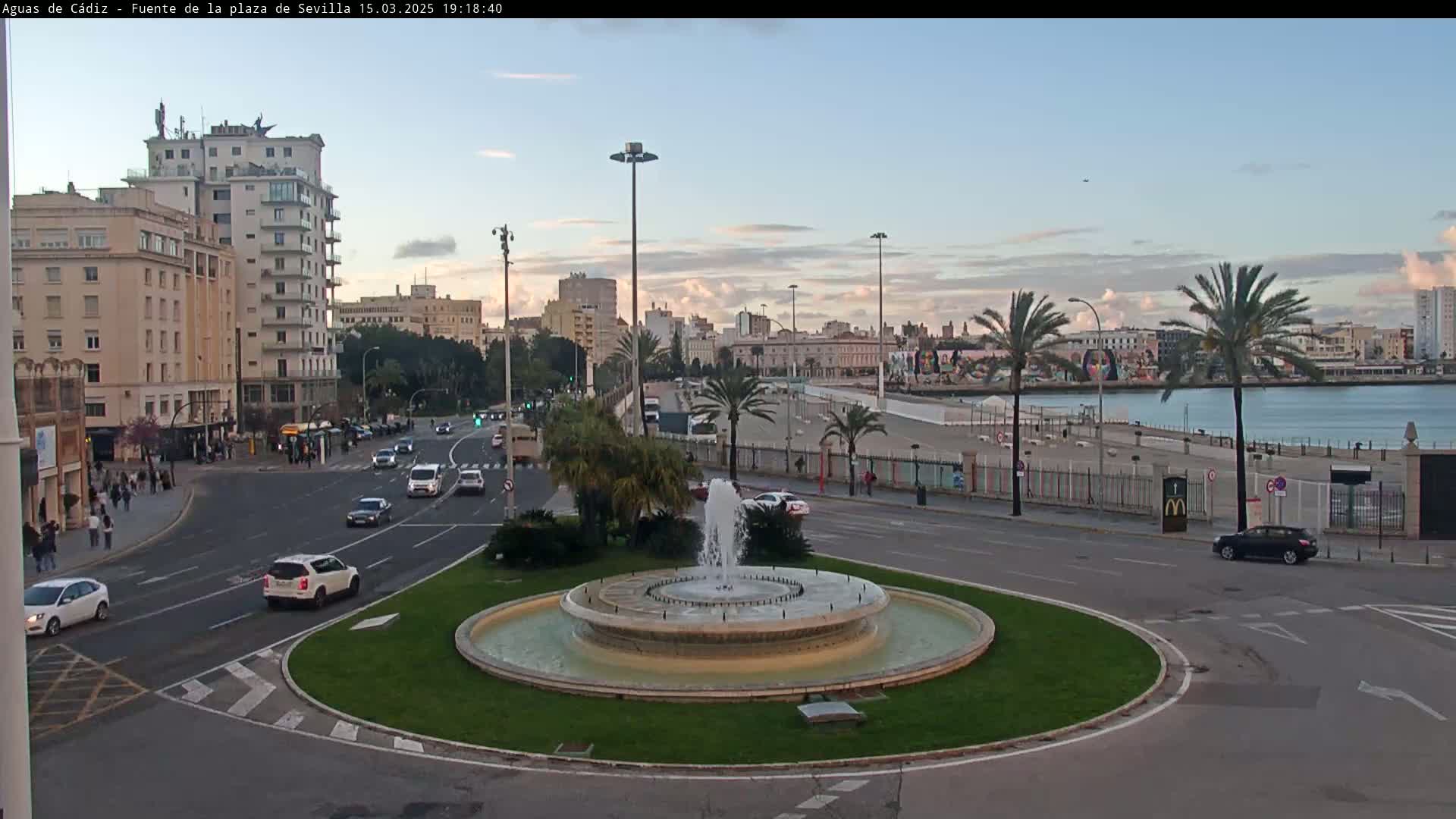A city waterfront scene at dusk shows a traffic circle with a fountain in the foreground, several buildings and palm trees in the middle ground, and a body of water in the background under a partly cloudy sky.