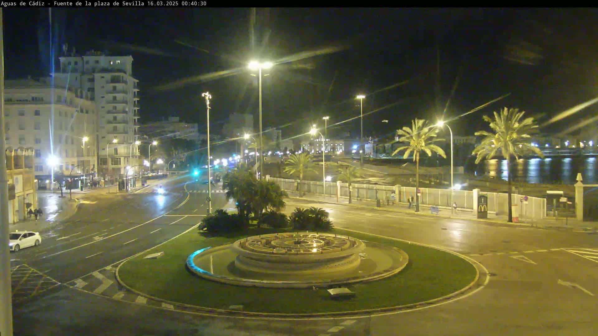 A nighttime view of a city square with a circular fountain in the center, wet roads, and palm trees lining a waterfront in the background.