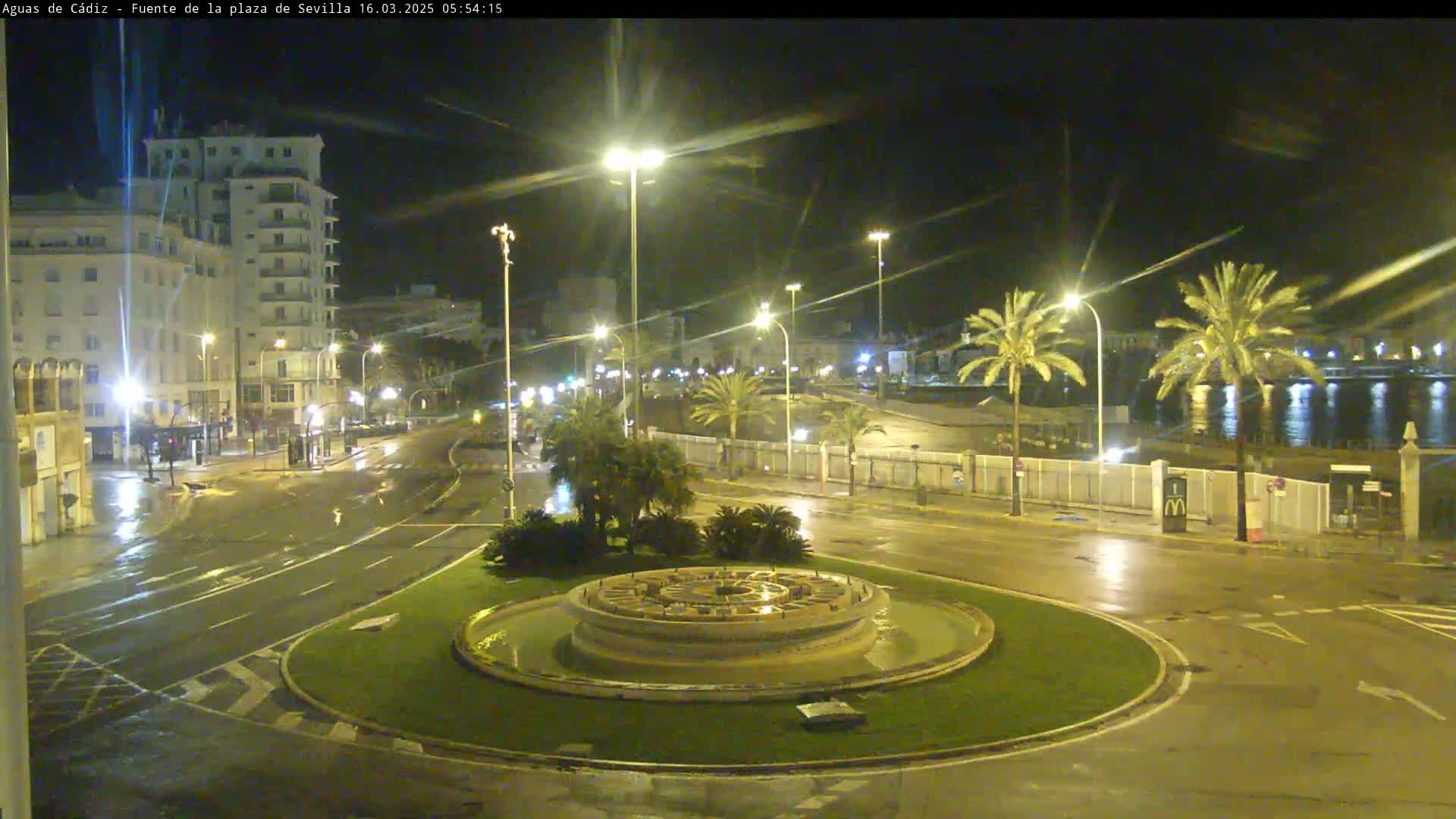 A nighttime view of a circular fountain in a plaza, surrounded by wet streets, palm trees, and buildings under bright streetlights in rainy weather.