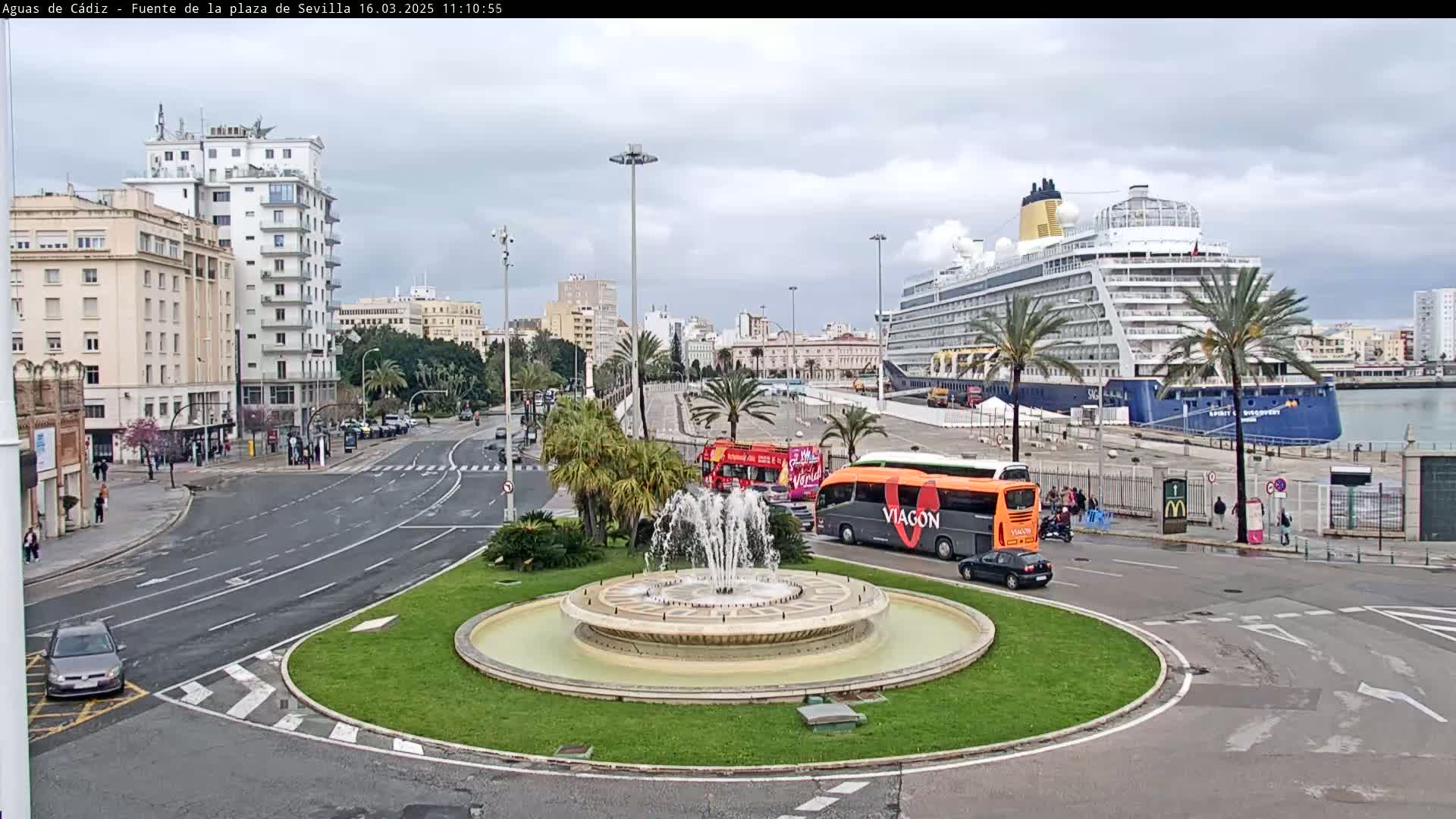 A city roundabout with a fountain in the center features a cruise ship docked in the harbor under an overcast sky, with buses and cars on the surrounding roads.