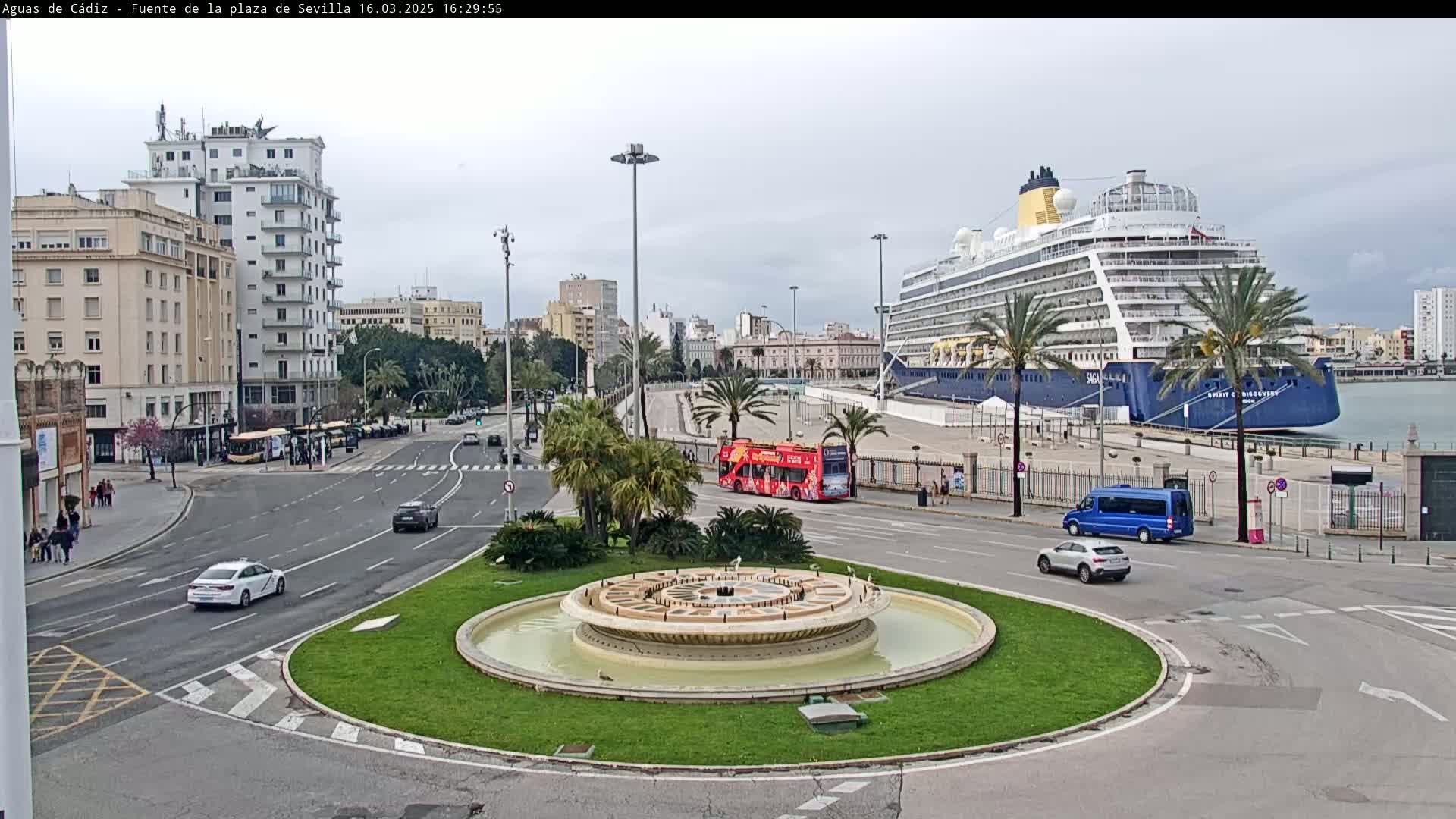 A large cruise ship is docked at a port next to a city with buildings, palm trees, a fountain, and several vehicles on the roads.  The weather is overcast.