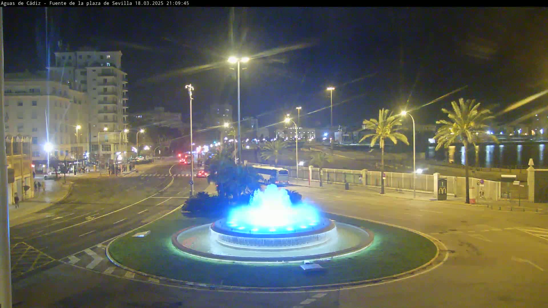 A nighttime view of a city square with a circular fountain illuminated in blue, surrounded by roads and buildings under clear skies.