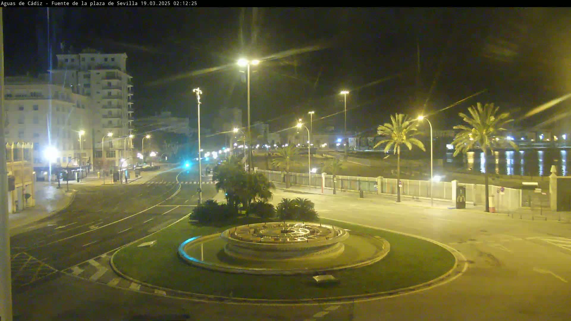 A nighttime view of a circular fountain in a city square, bordered by palm trees, buildings, and a roadway, near a body of calm water.
