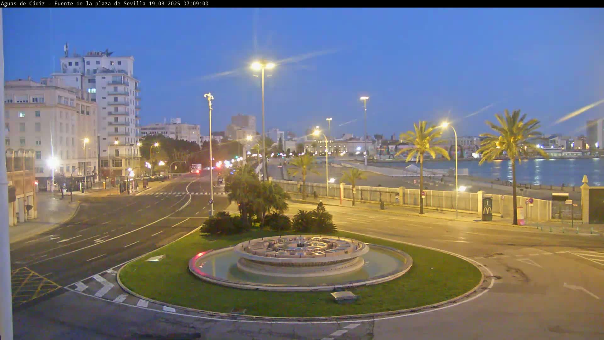 A circular fountain sits in a city roundabout at twilight, with palm trees and a waterfront visible in the background under a clear sky.