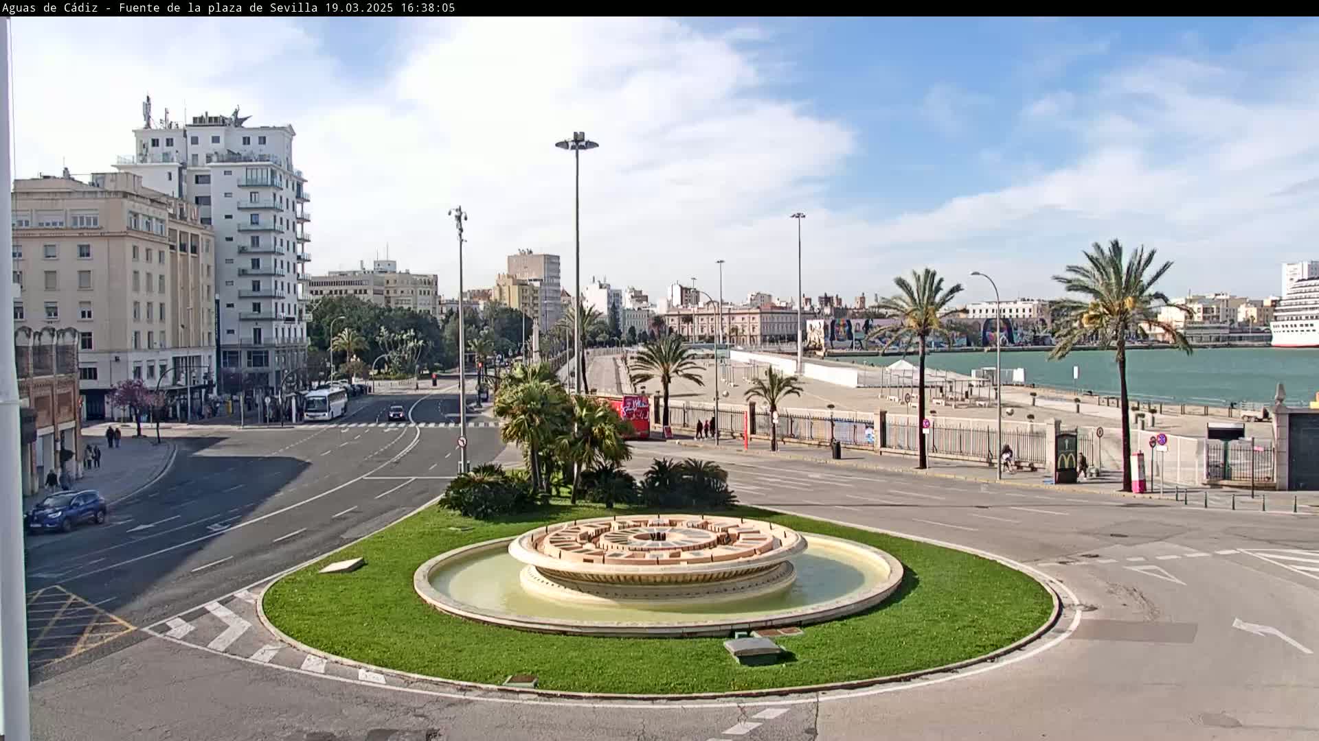 A circular fountain sits in a city plaza, with palm trees lining a waterfront promenade beyond a roadway under a partly cloudy sky.