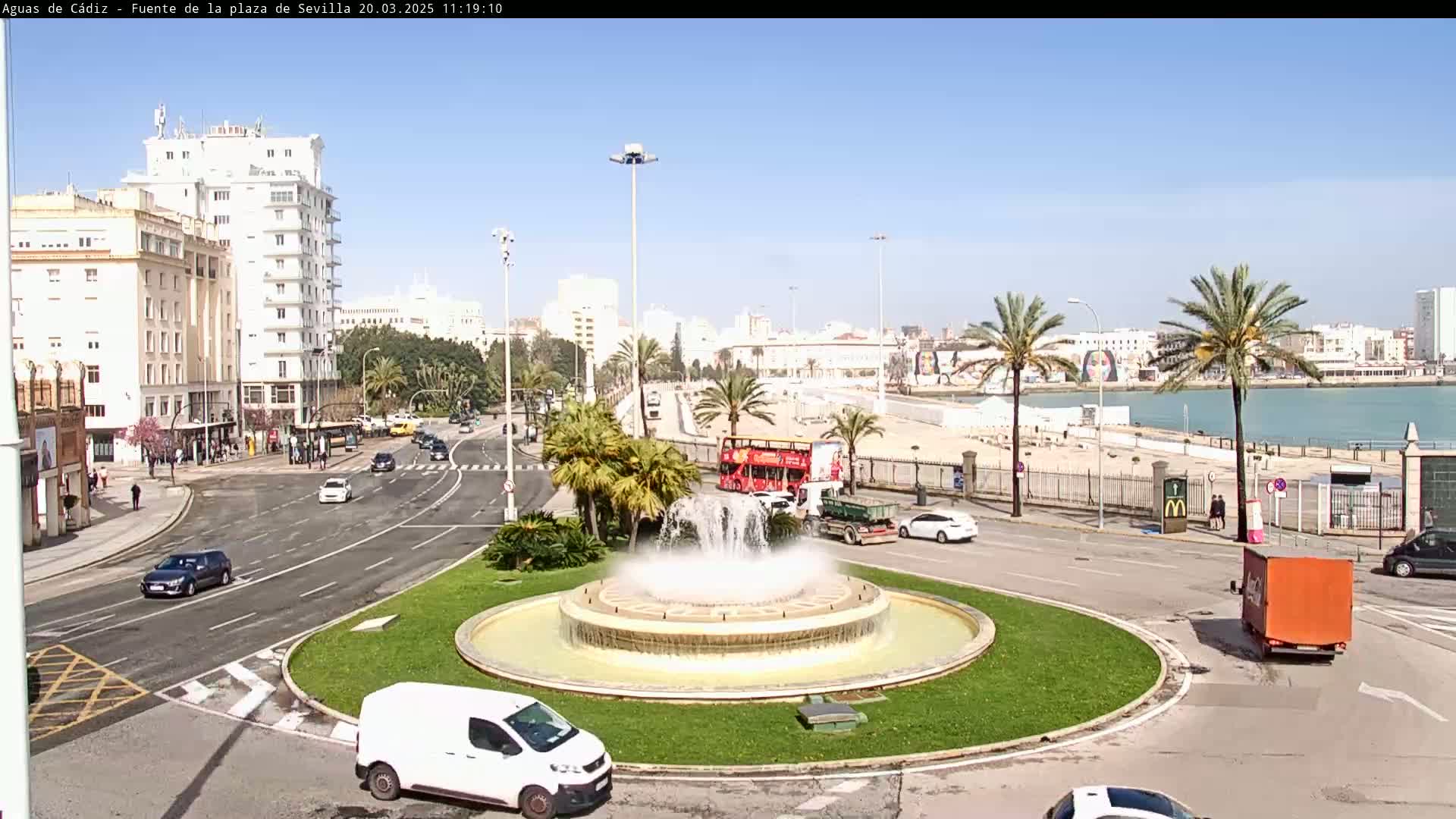 A city roundabout with a fountain at its center, surrounded by palm trees, buildings, and cars, under a clear sunny sky.