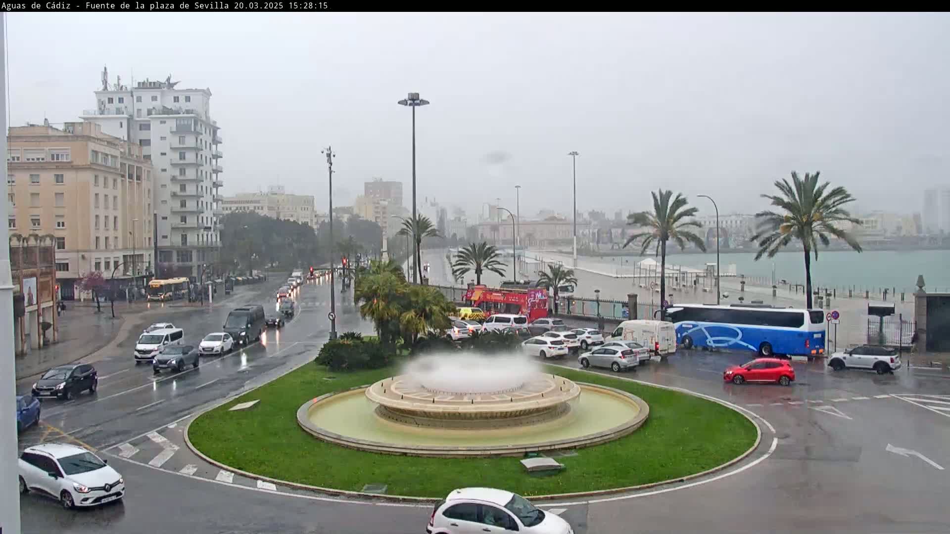A city roundabout with a fountain in the center is surrounded by traffic on a rainy day, with palm trees and buildings visible in the background near a body of water.