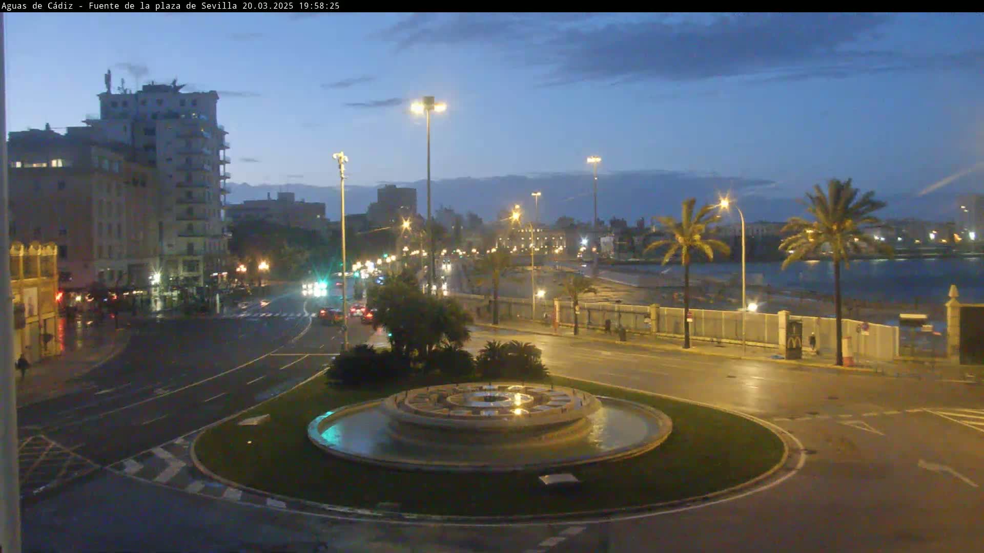A circular fountain sits in a traffic circle at dusk, near a shoreline with palm trees and buildings under a twilight sky.