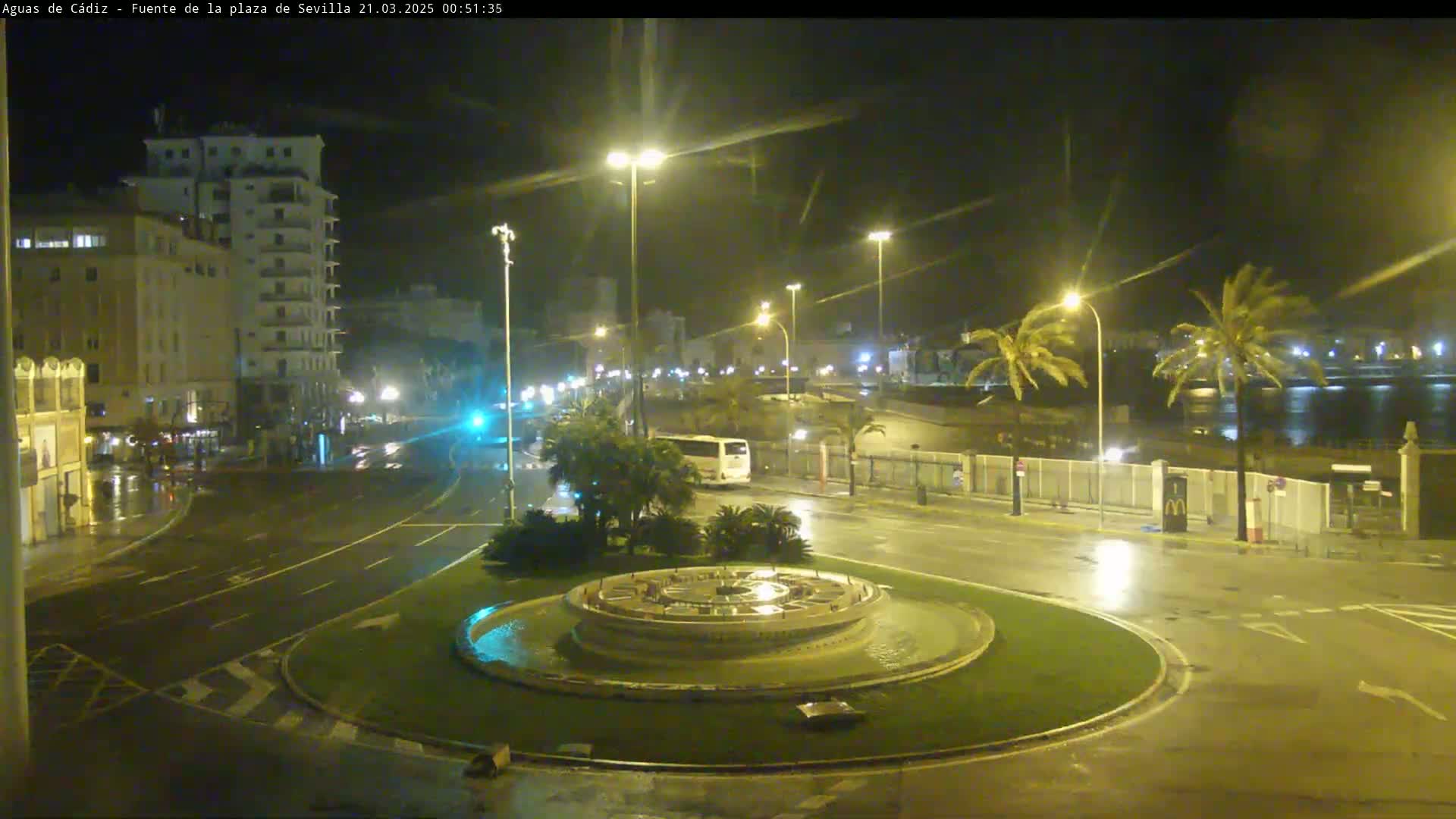 A nighttime, wet scene shows a circular fountain in the center of a plaza, surrounded by streets, buildings, and palm trees under streetlights.