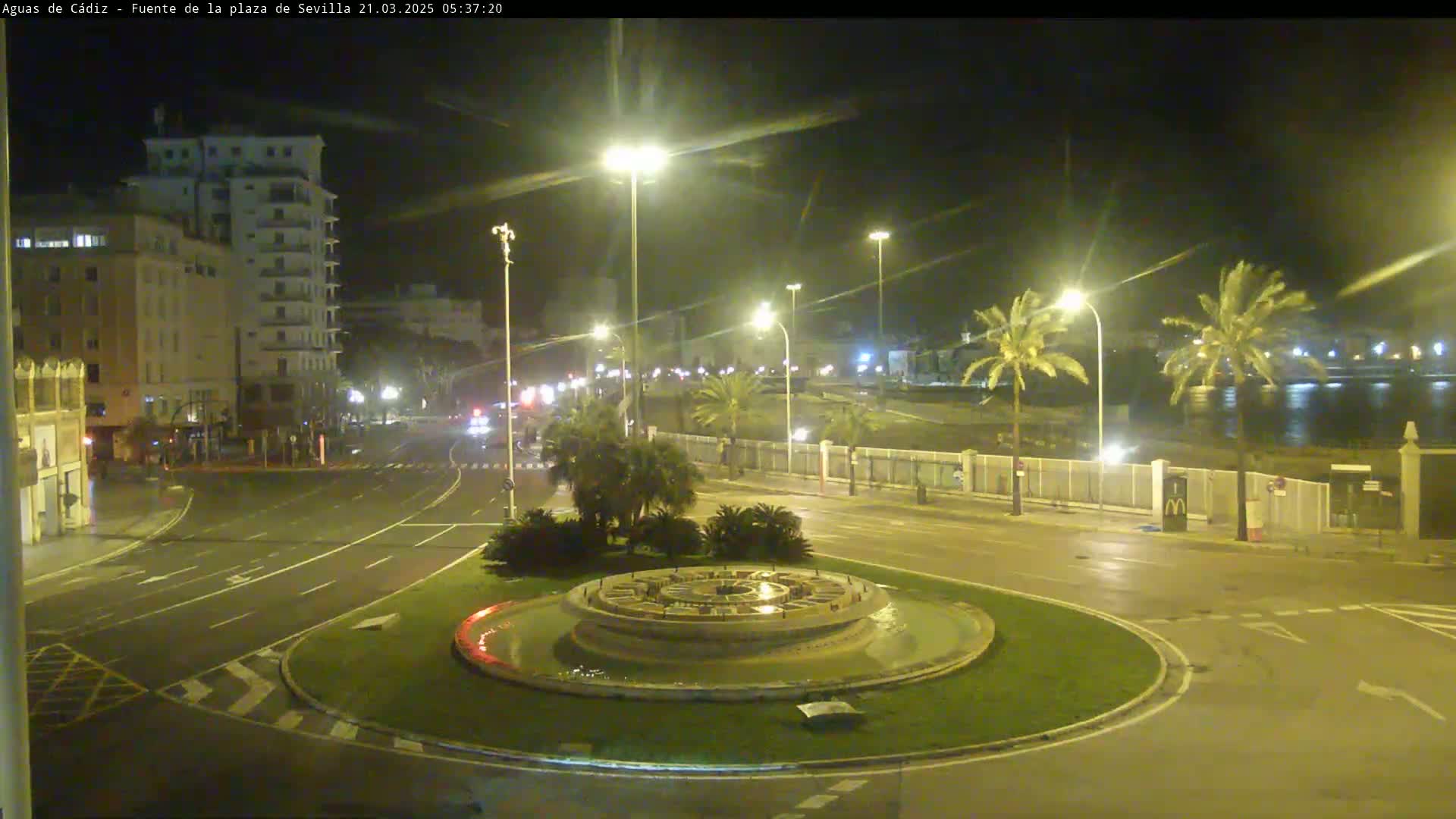A nighttime view of a circular fountain in a city square, with wet roads and palm trees lining a waterfront in the background under slightly overcast skies.