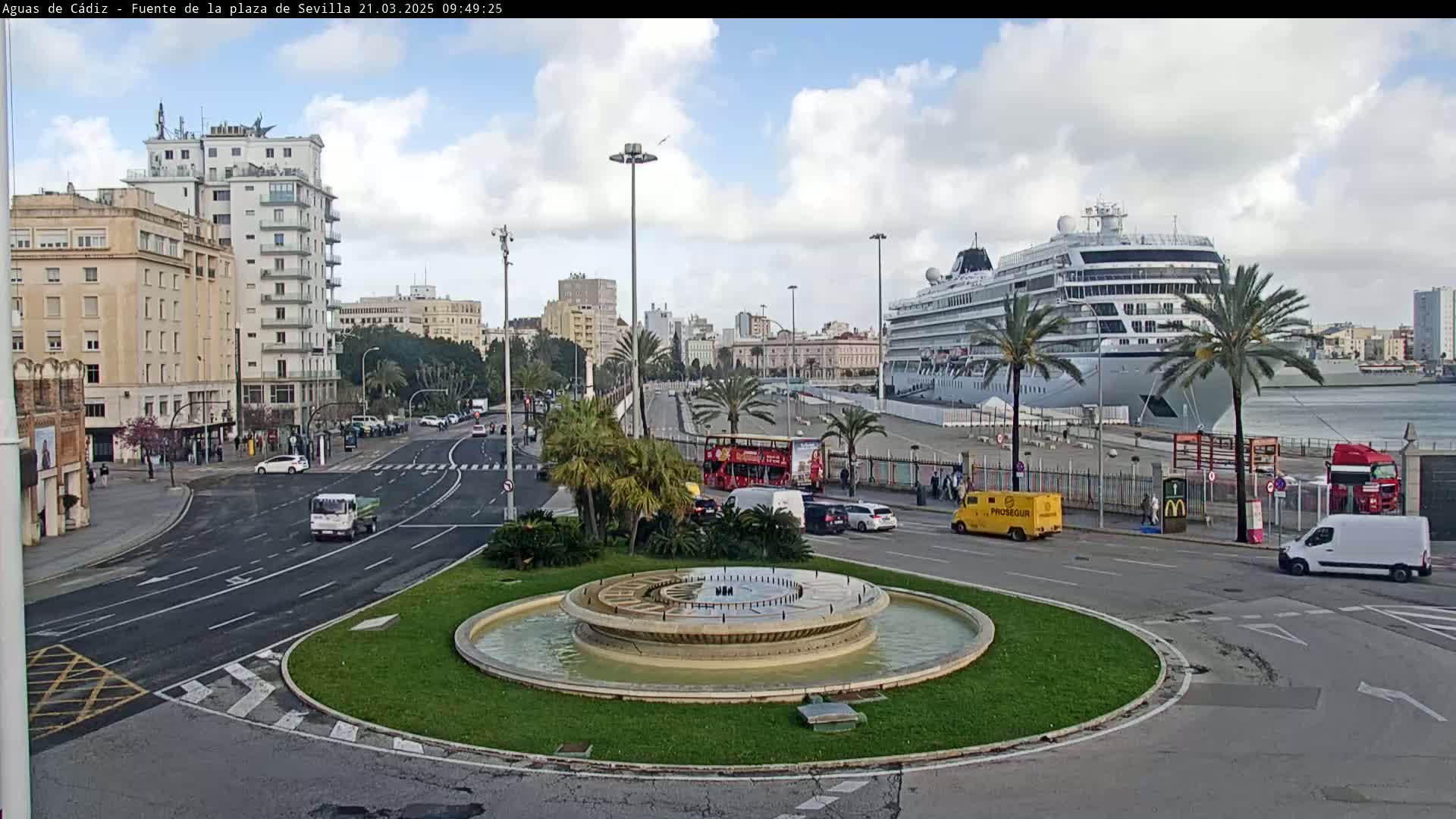A city roundabout with a fountain at its center, featuring a large cruise ship docked at the harbor in the background under a partly cloudy sky.