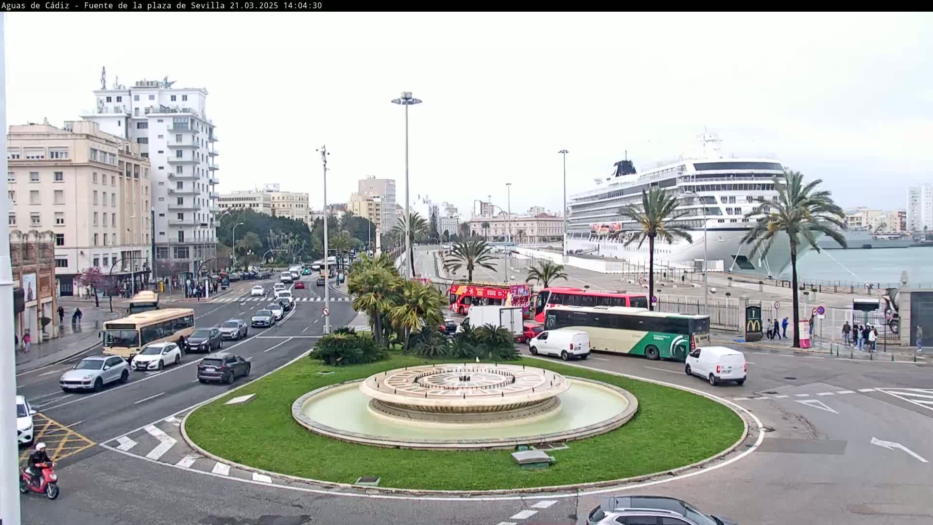 A city roundabout with a central fountain is shown, with a large cruise ship docked in the harbor in the background on an overcast day.