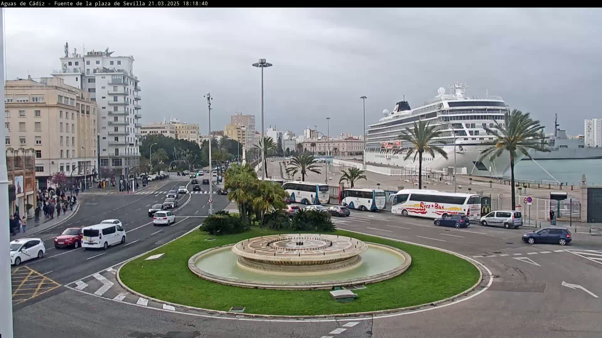 A large cruise ship is docked at a port, near a roundabout with a fountain and several cars and buses, under an overcast sky.