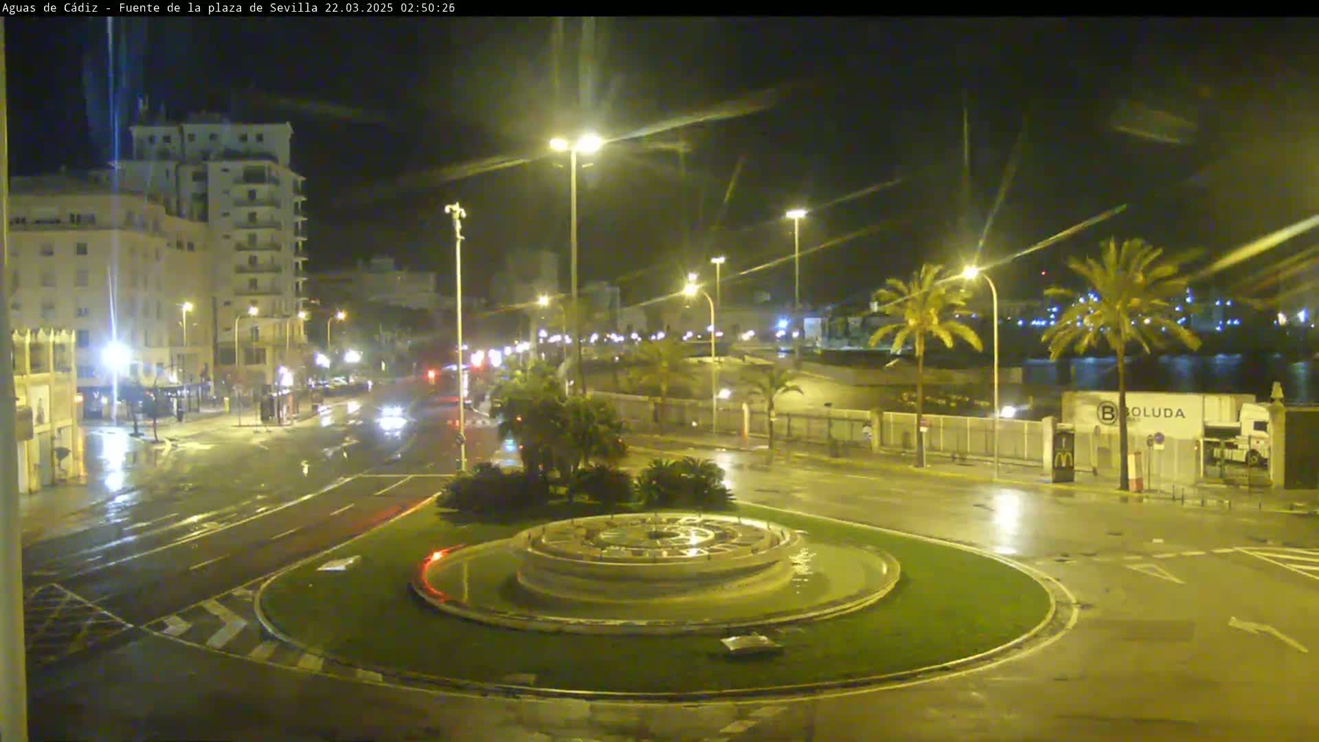 A nighttime view of a wet city roundabout with a circular fountain in the center, surrounded by palm trees, buildings, and streetlights, under a dark sky.