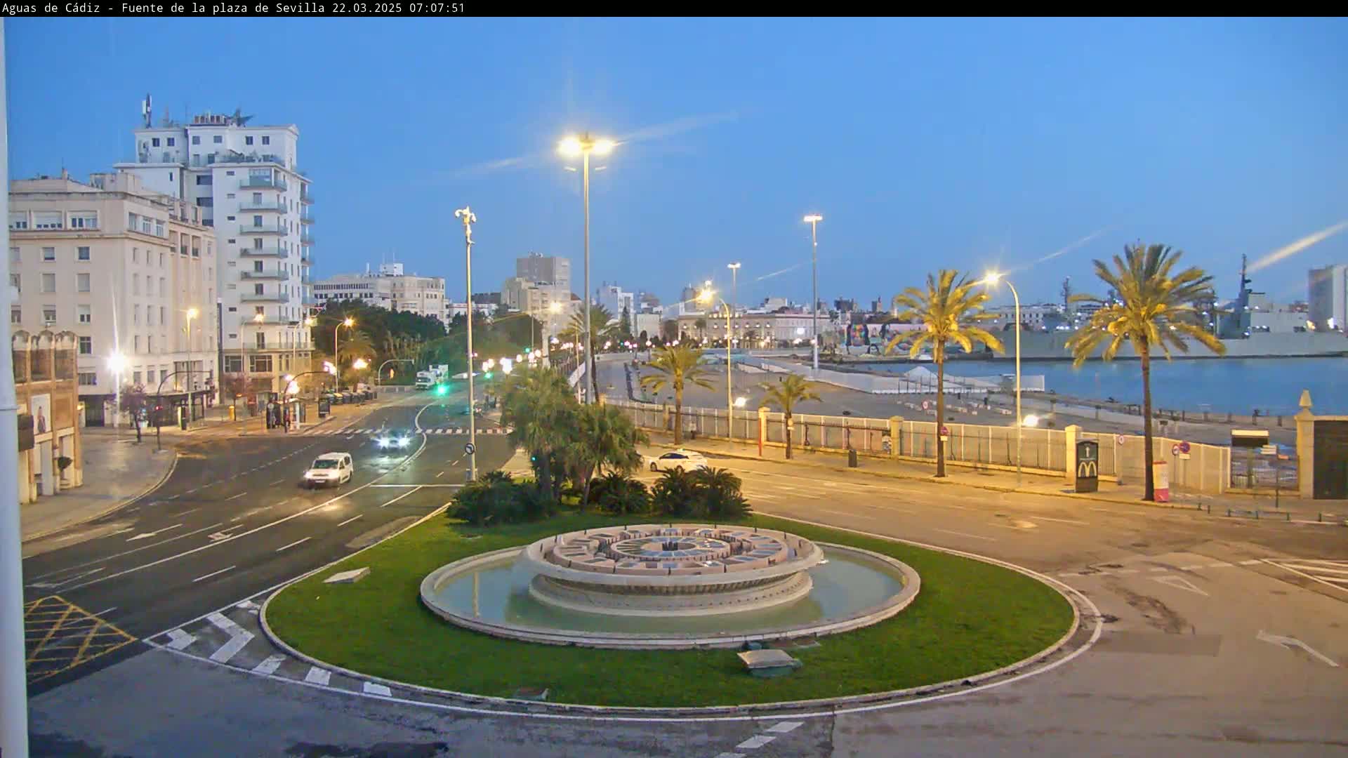 A circular fountain sits in a traffic circle at twilight near a harbor with palm trees and several buildings under a clear, dusky sky.