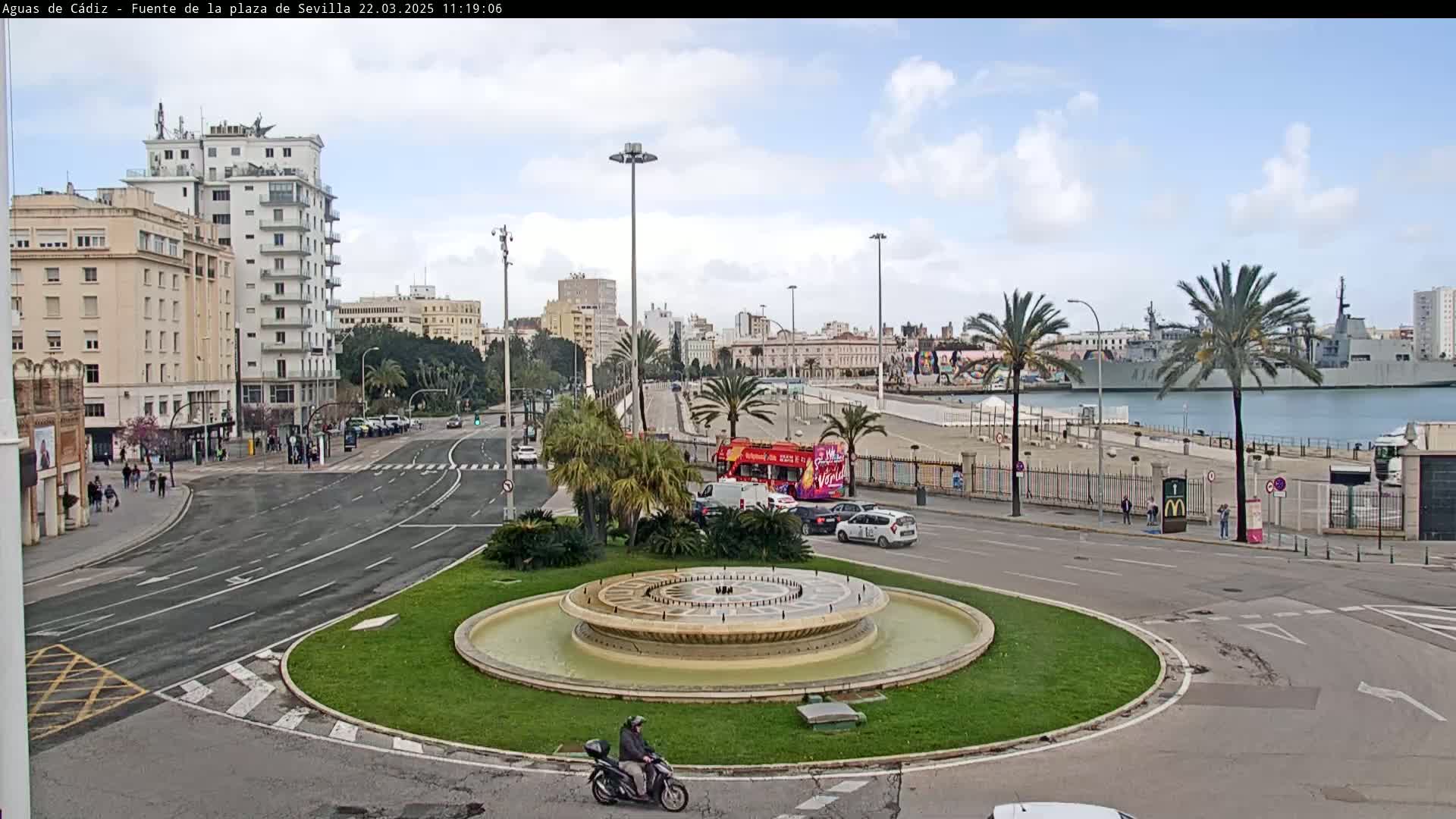 A circular fountain sits in a traffic circle near a waterfront, with buildings and palm trees visible under a partly cloudy sky.