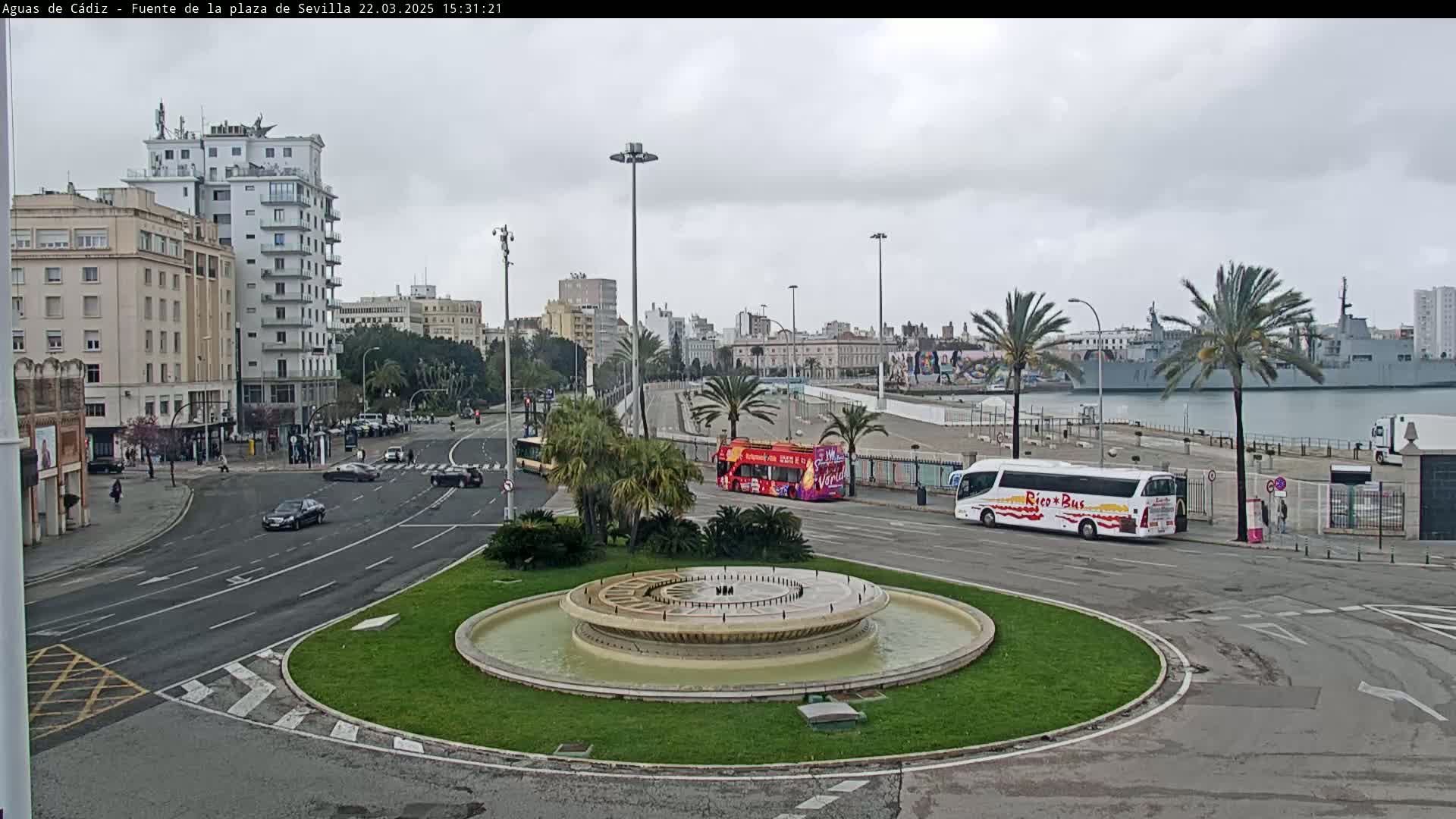A city street scene under an overcast sky features a circular fountain in the foreground, with several cars and two tour buses on a roadway near a harbor where a large ship is docked.