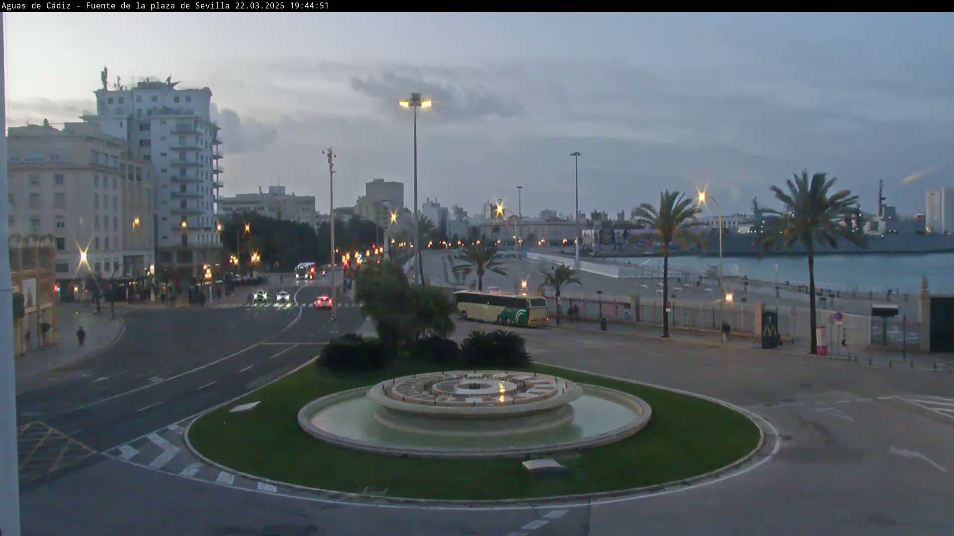 A city roundabout with a circular fountain in the center, streetlights illuminating the area at dusk, overlooks a harbor with a large ship and palm trees lining the waterfront.