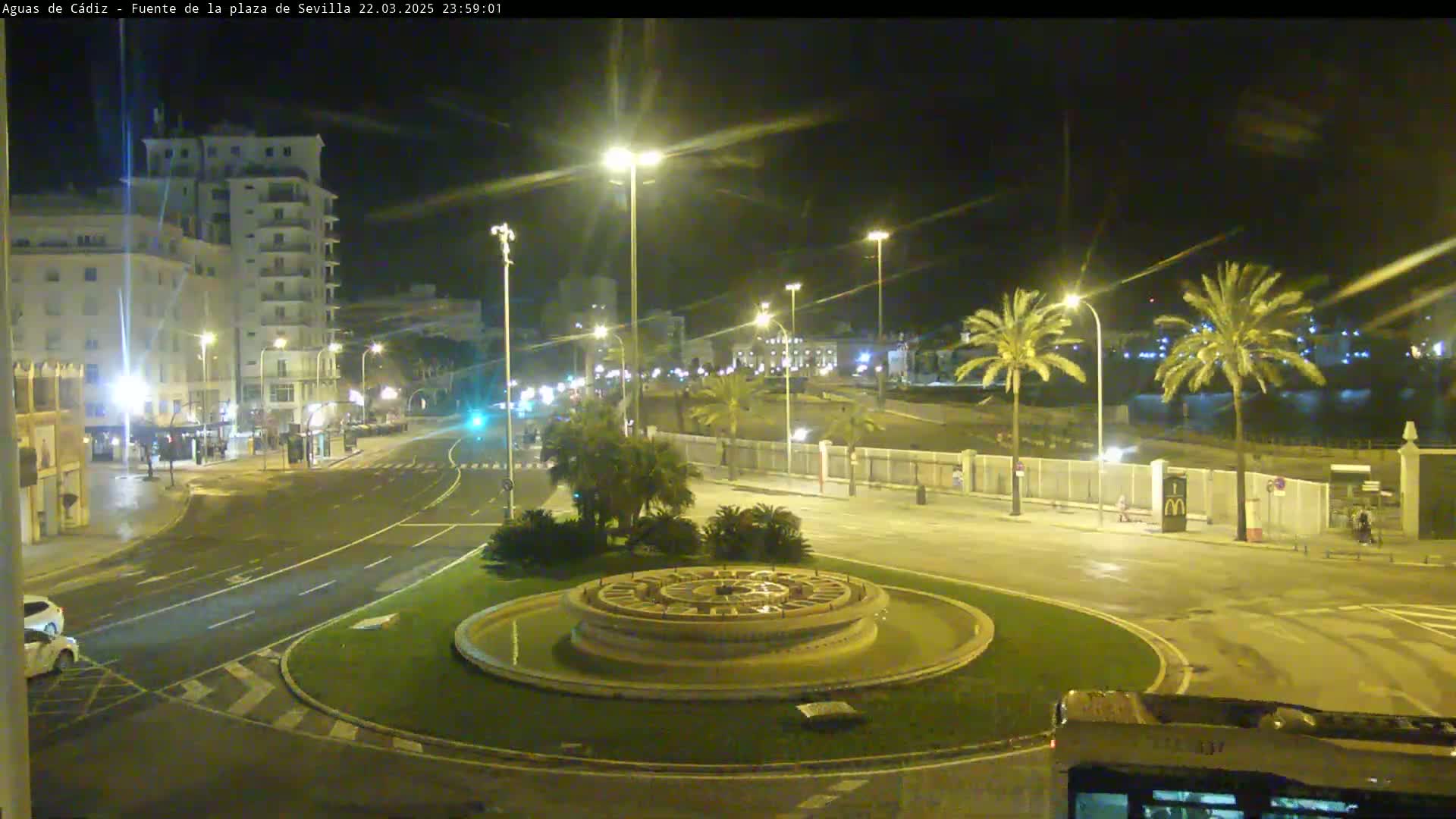 A nighttime view of a city roundabout with a circular fountain at its center, surrounded by palm trees, buildings, and streetlights.