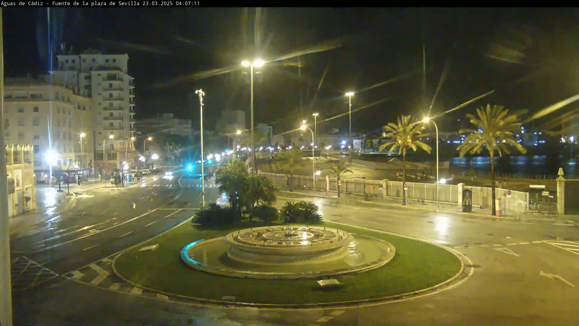 A nighttime, rain-slicked roundabout with a circular fountain in the center, surrounded by palm trees and buildings under streetlights.
