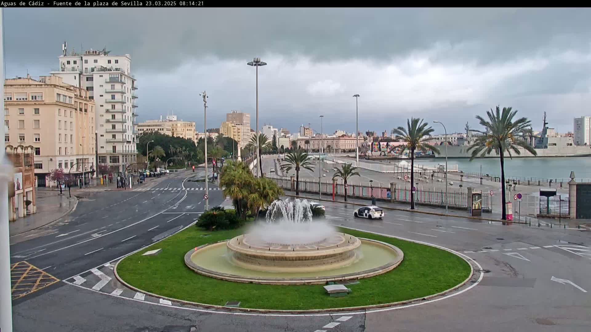 A city square with a fountain in the center, surrounded by roads and buildings, overlooks a harbor with a ship under a cloudy sky.