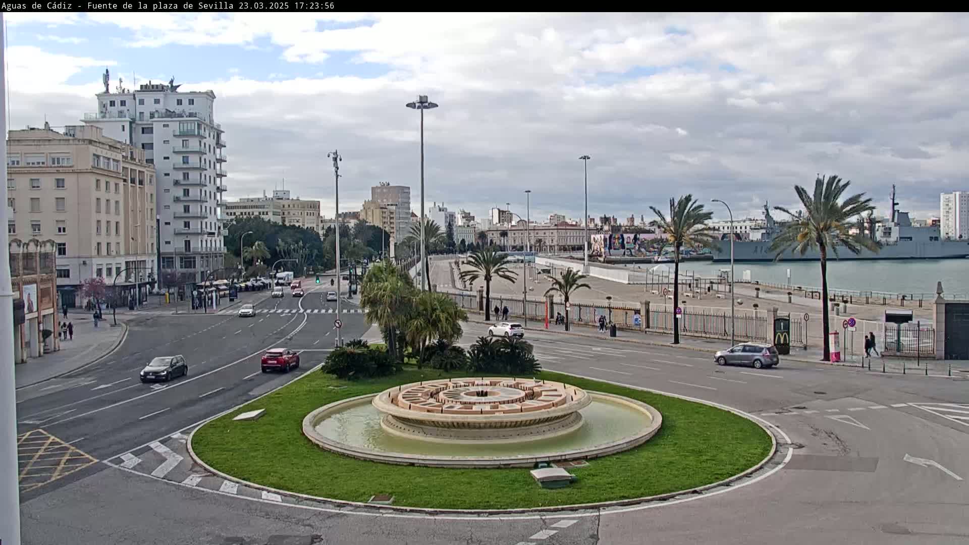 A circular fountain sits in the center of a traffic circle, with city buildings, palm trees, and a docked warship visible in the background under a mostly cloudy sky.