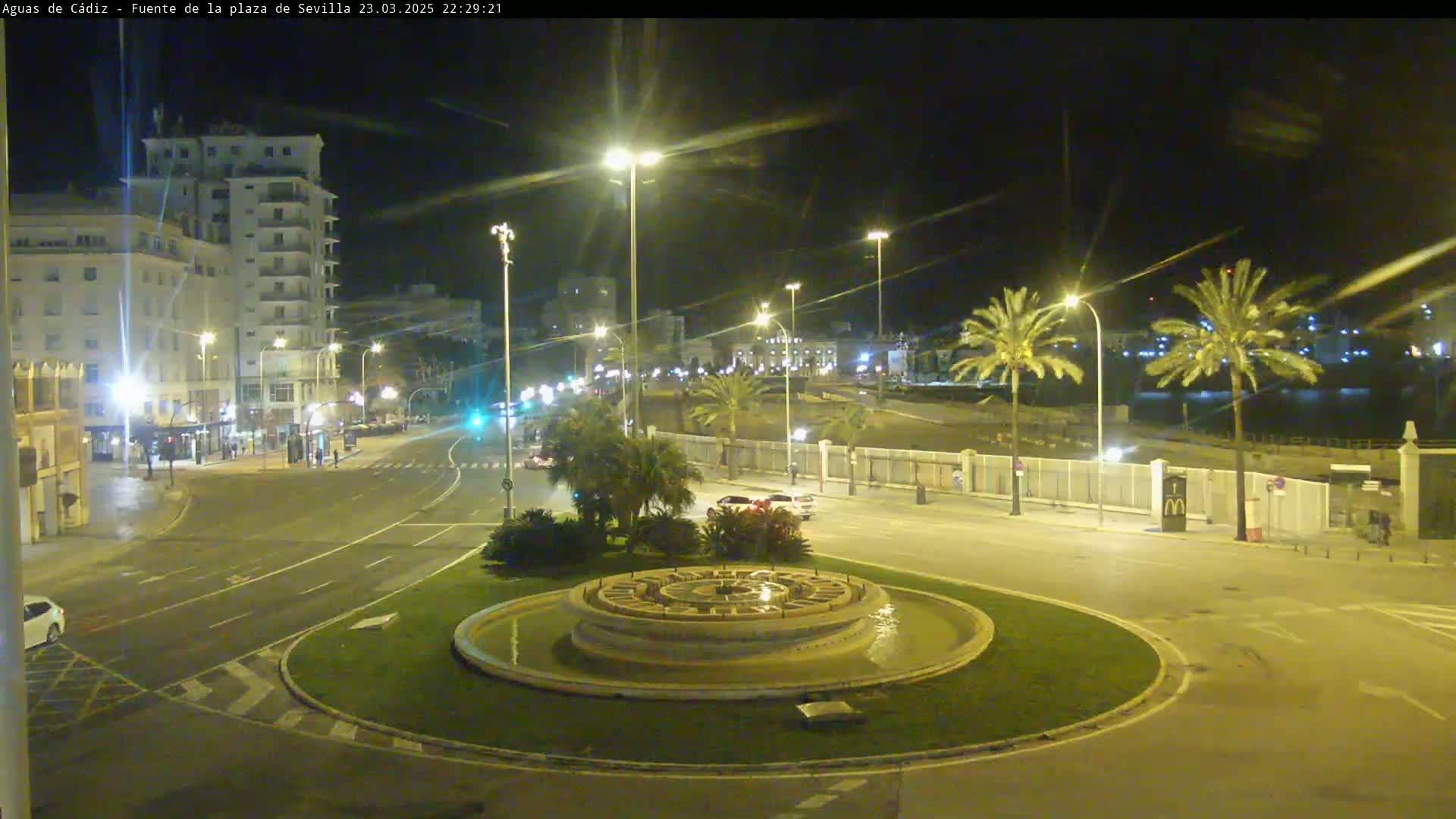 A nighttime view of a city roundabout with a circular fountain in the center, surrounded by palm trees, buildings, and sparsely trafficked roads.
