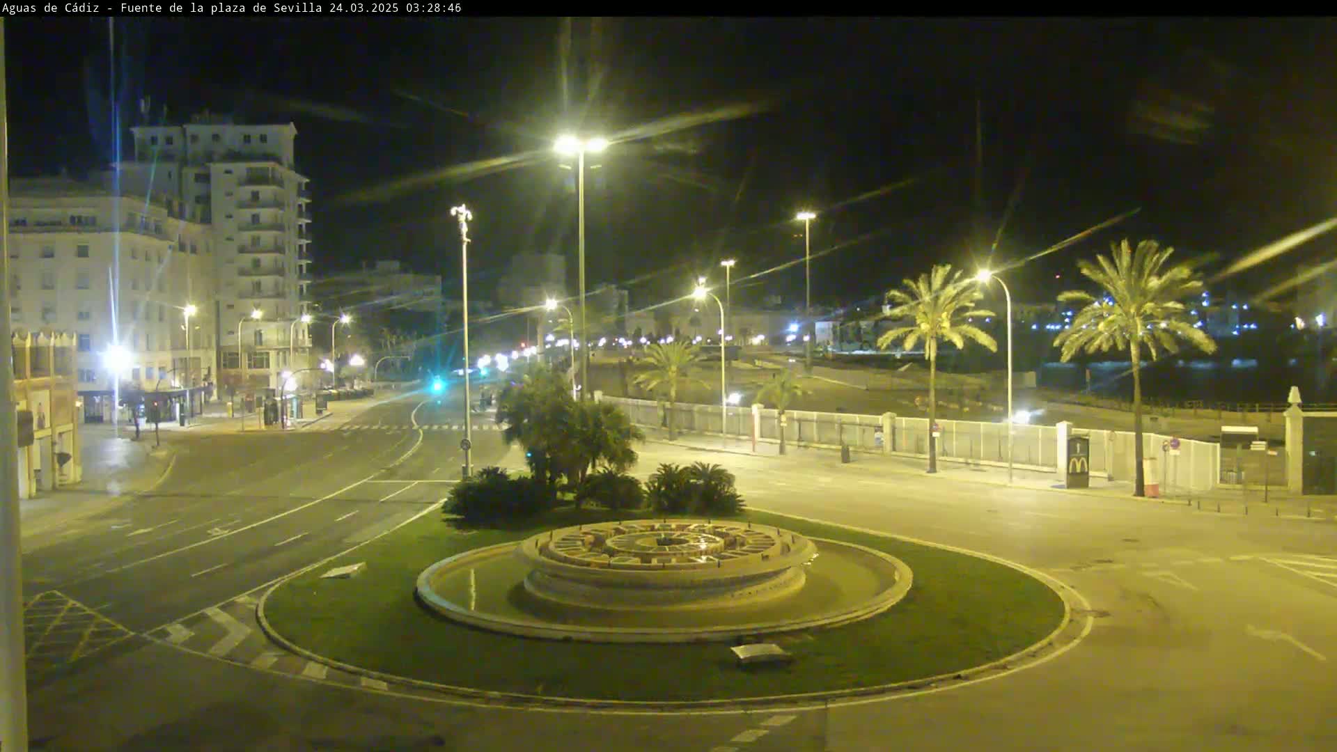 A nighttime view of a circular fountain in a city square, surrounded by palm trees, buildings, and roadways illuminated by streetlights.