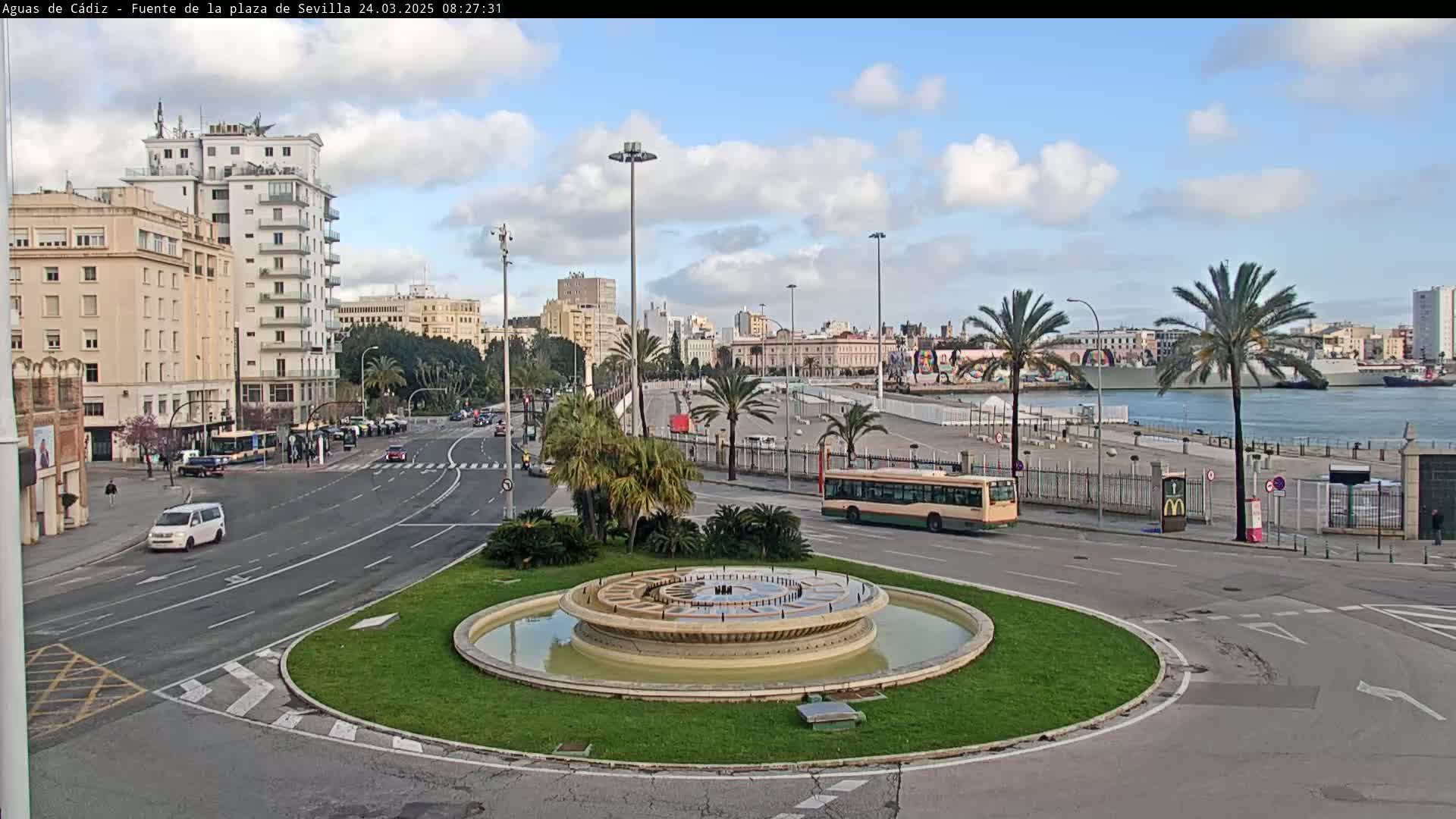 A circular fountain sits in the center of a traffic circle, with city buildings, palm trees, a bus, and a harbor with docked ships visible in the background under a partly cloudy sky.