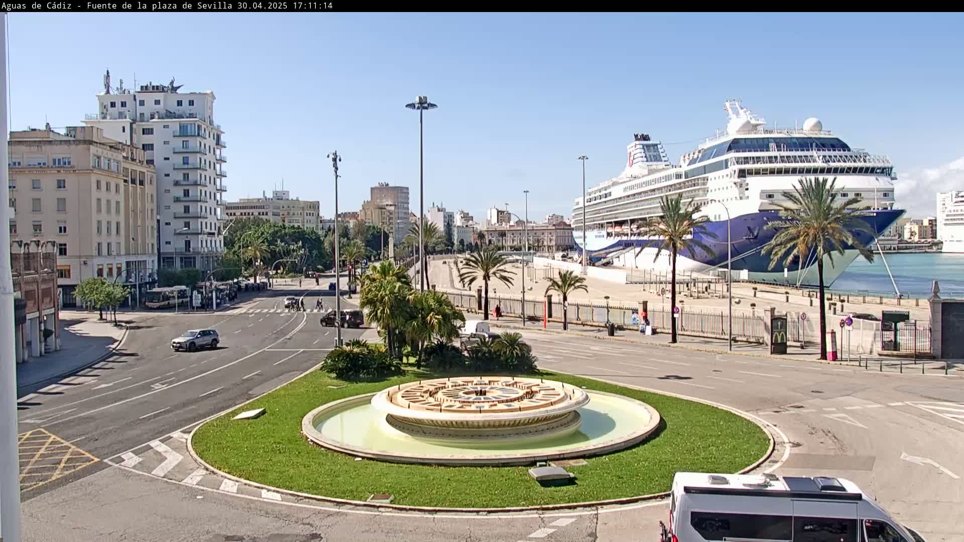 A large cruise ship is docked at a port next to a city featuring a circular fountain in the middle of a road intersection, under a clear sunny sky.