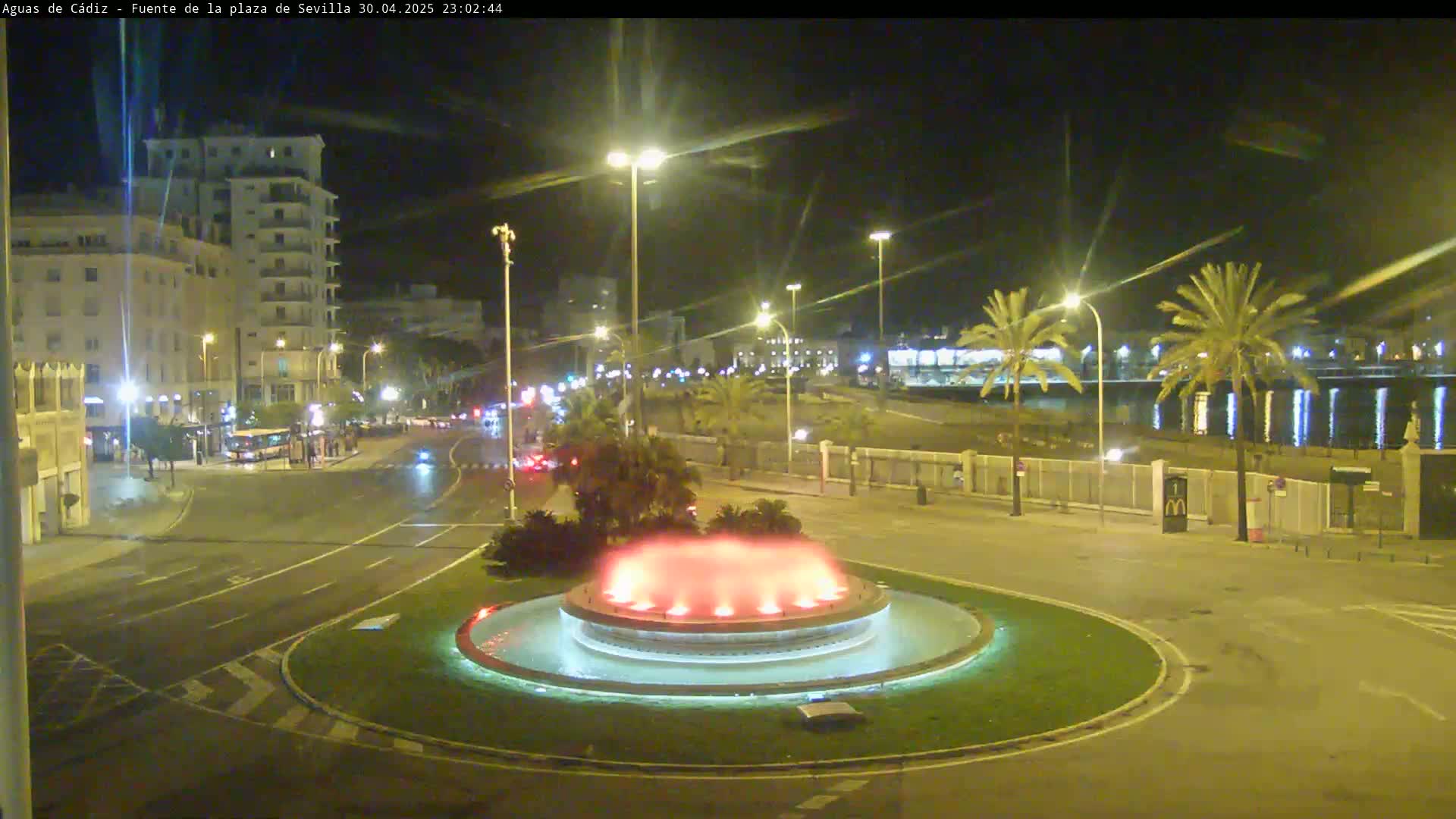 A nighttime view of a city square featuring a circular fountain with red lights, illuminated buildings, palm trees, and a calm body of water in the background.