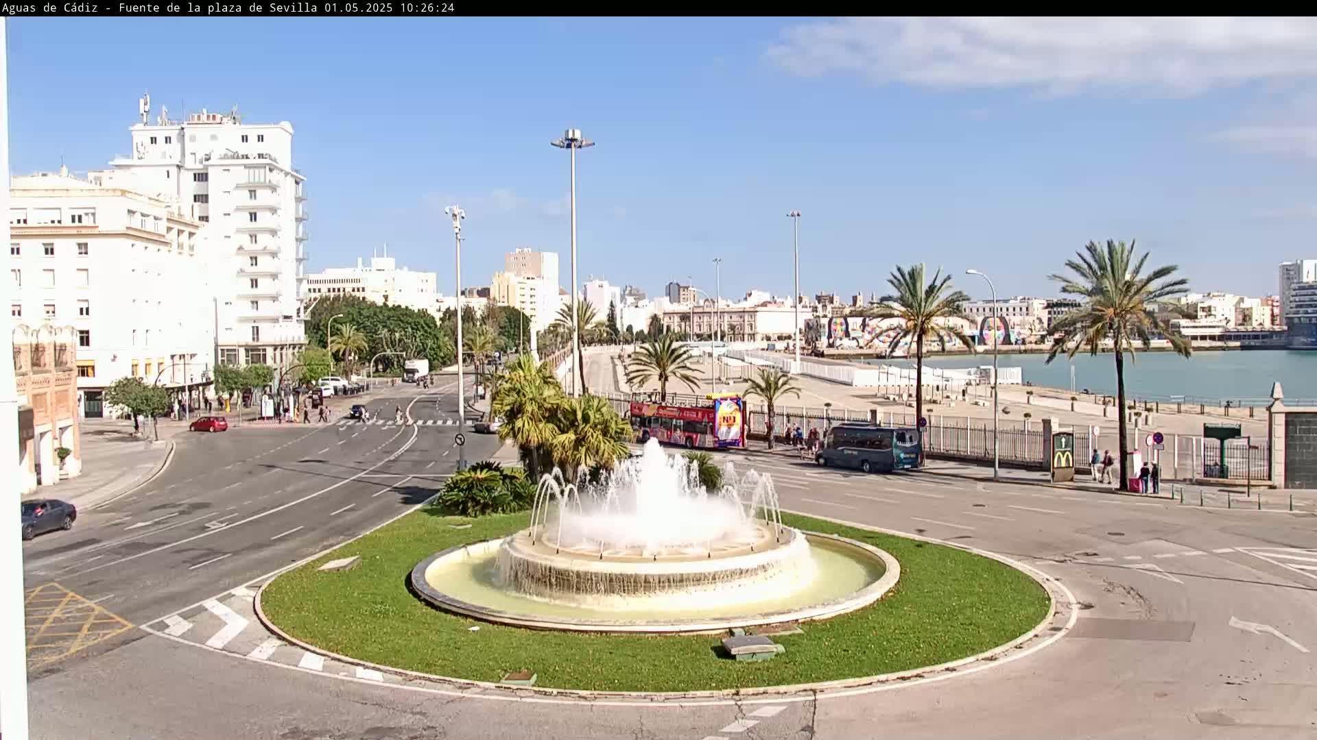 A circular fountain sprays water in a plaza near a waterfront, with palm trees and buildings lining a sunny street.