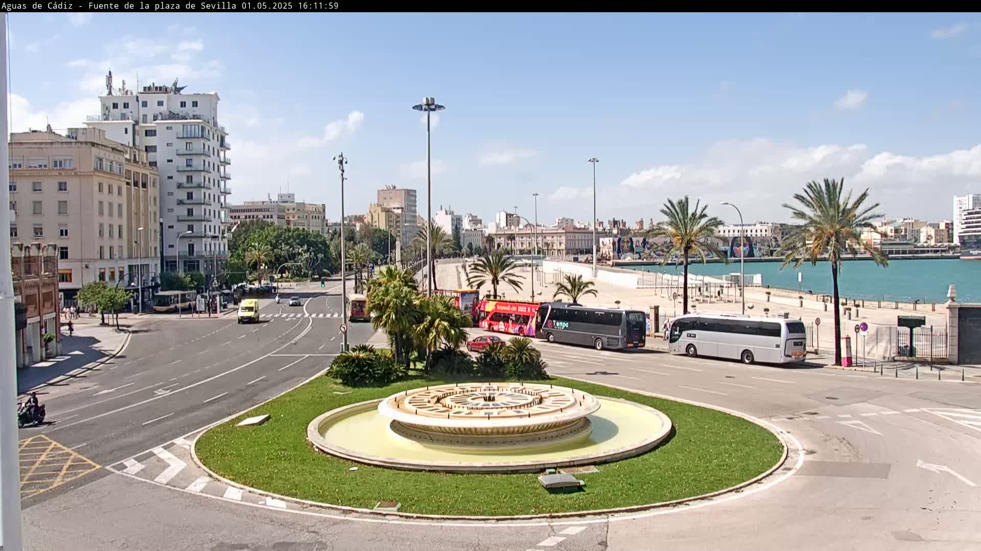 A circular fountain sits in a traffic circle near a waterfront, with several buses and other vehicles on a roadway lined with palm trees under a partly cloudy sky.