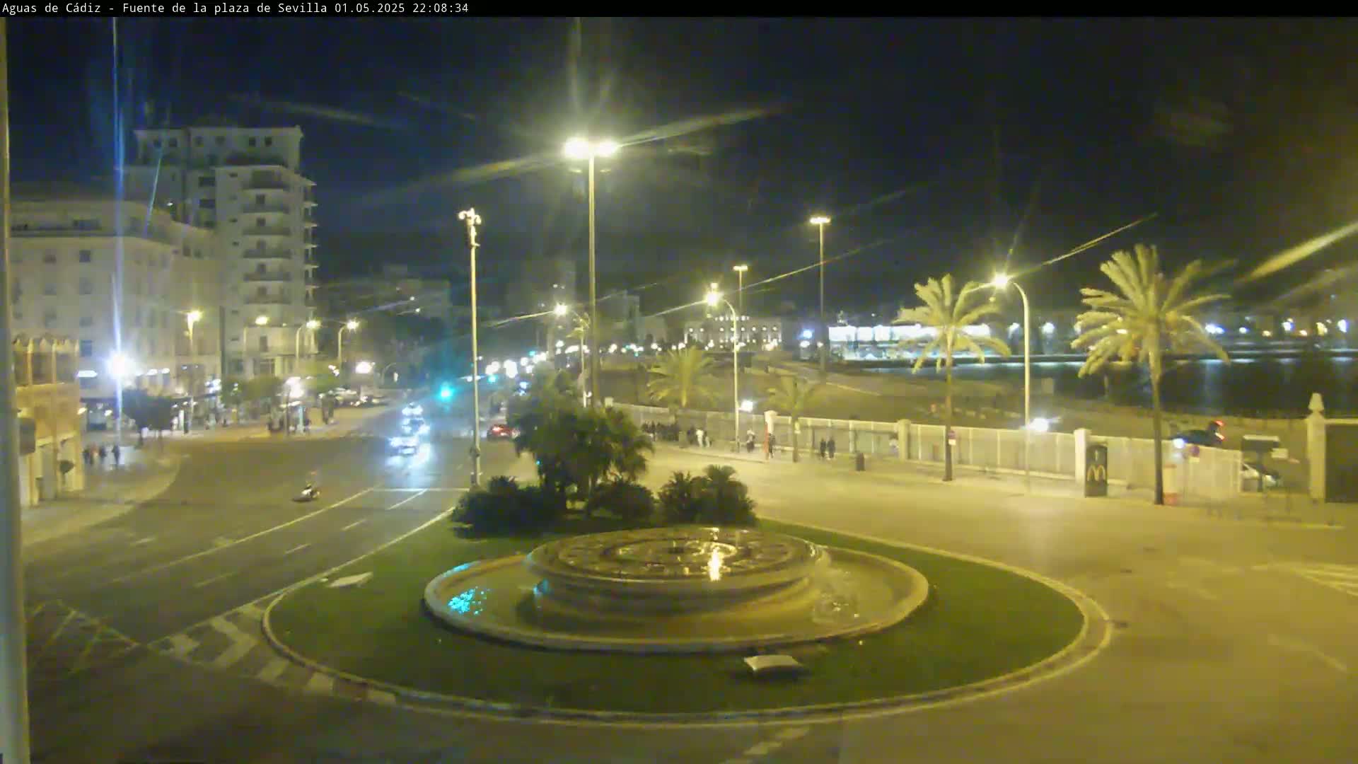 A nighttime view of a city square with a circular fountain in the center, palm trees lining a nearby waterfront, and streetlights illuminating the scene.