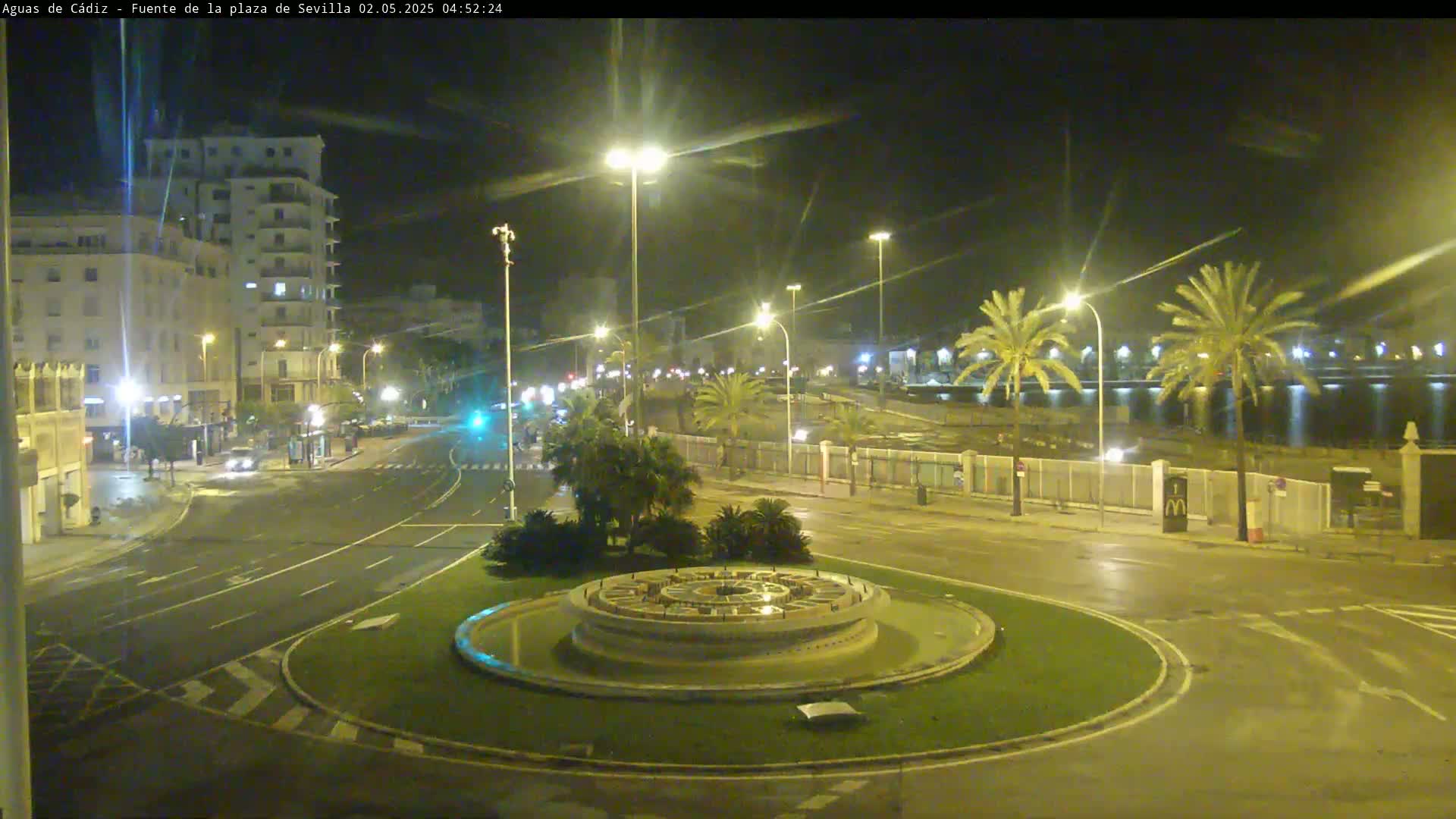 A nighttime view of a circular fountain in a plaza, surrounded by streets, buildings, and palm trees, under clear skies.