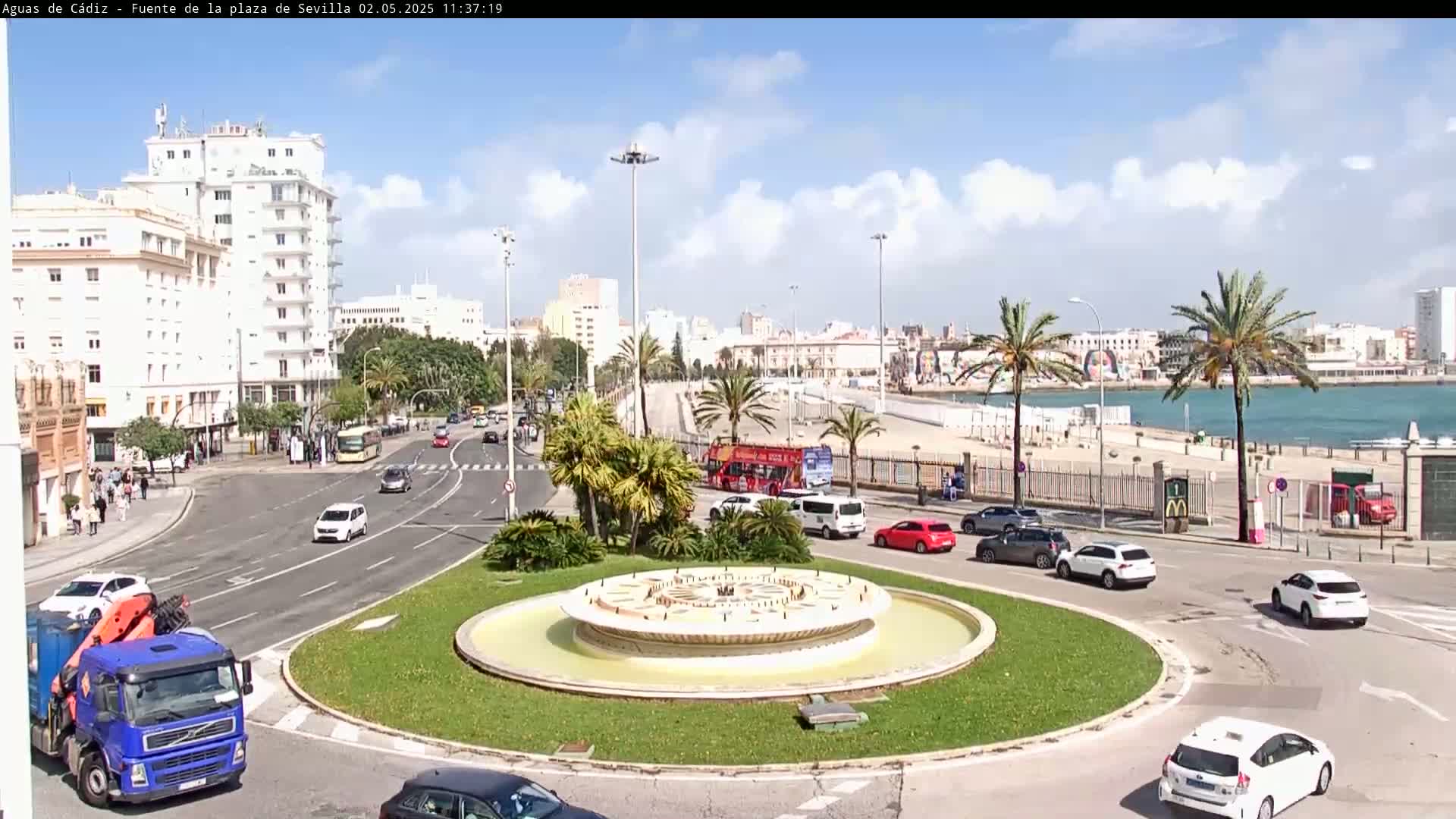 A traffic circle with a fountain in the center is surrounded by buildings, palm trees, cars, and a beach with a body of water beyond it under a partly cloudy sky.