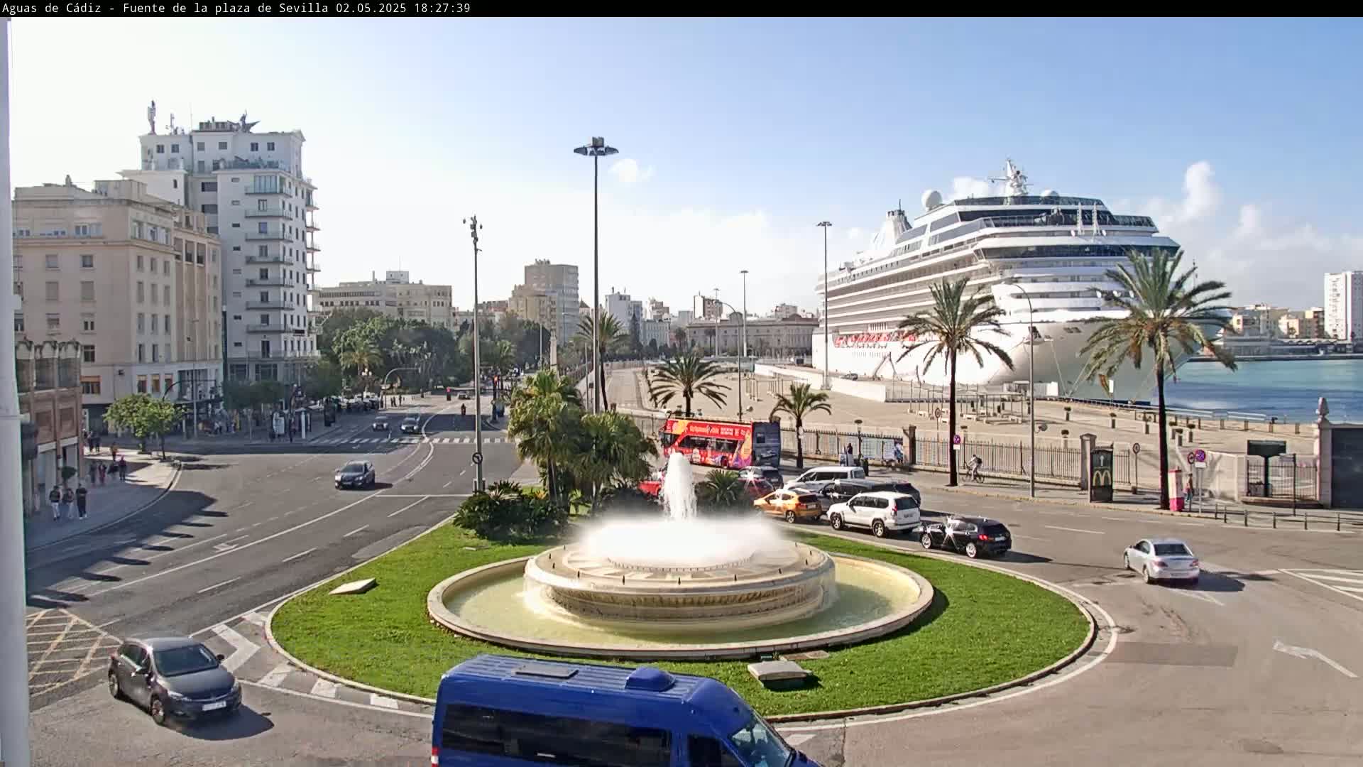 A large cruise ship is docked at a port near a city with a fountain in a traffic circle, under a mostly sunny sky.