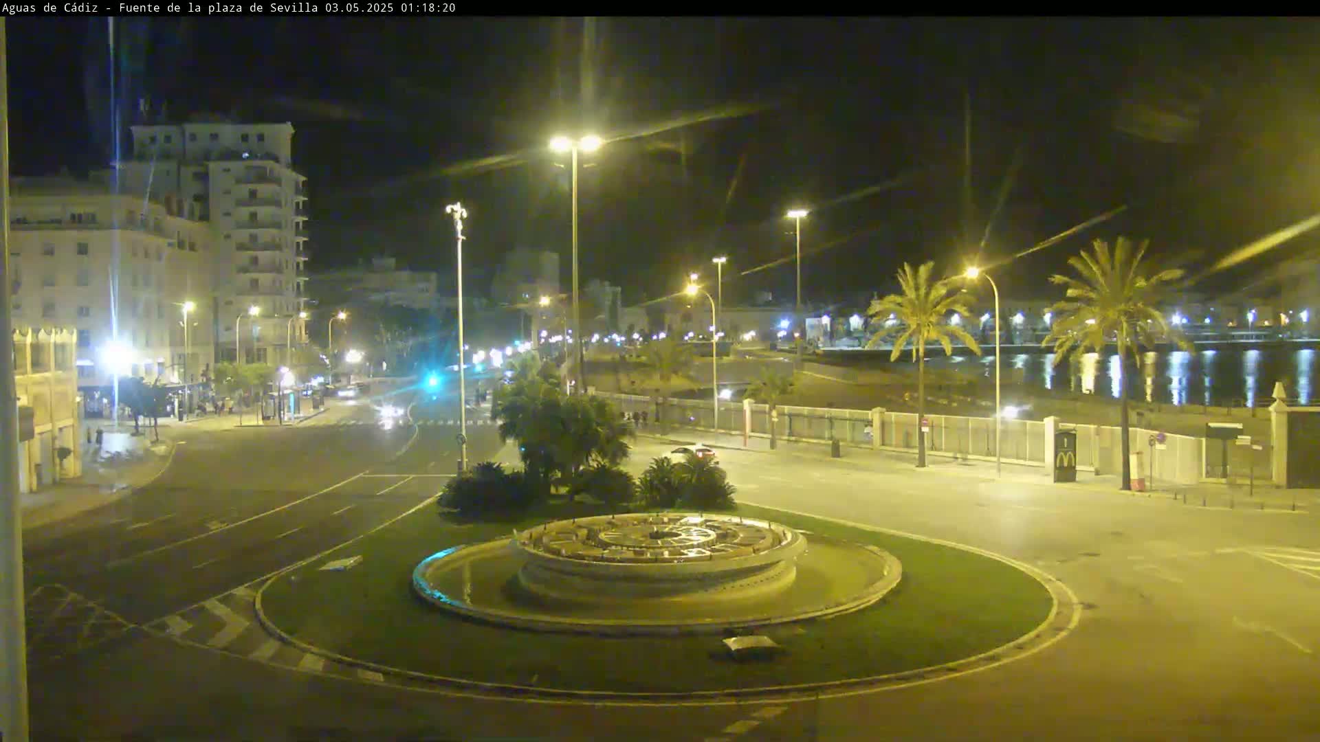 A nighttime view of a circular traffic island with a fountain in the center, surrounded by streets and buildings, near a body of water under clear skies.