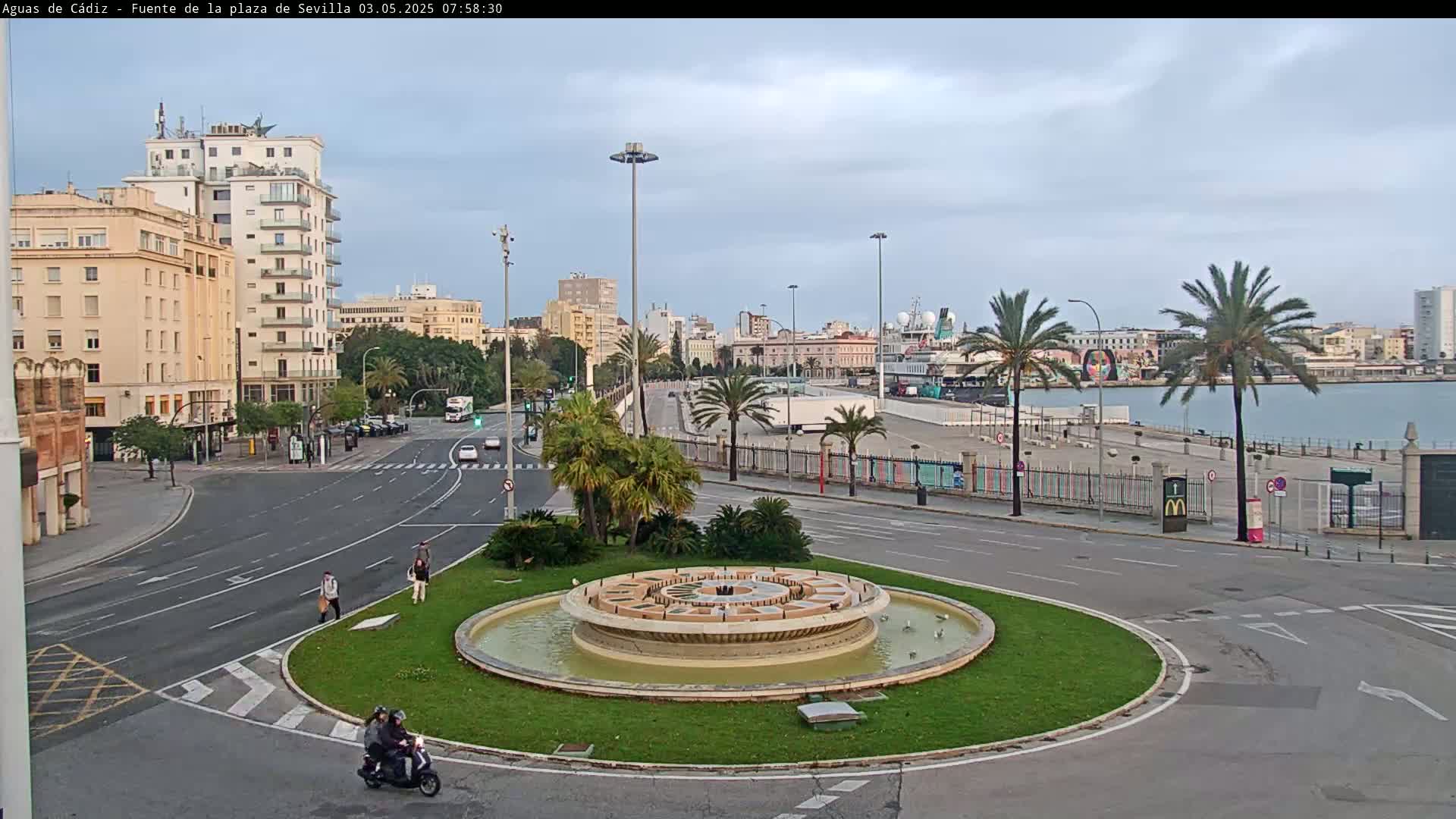 A circular fountain sits in a traffic circle overlooking a waterfront city under a mostly cloudy sky.