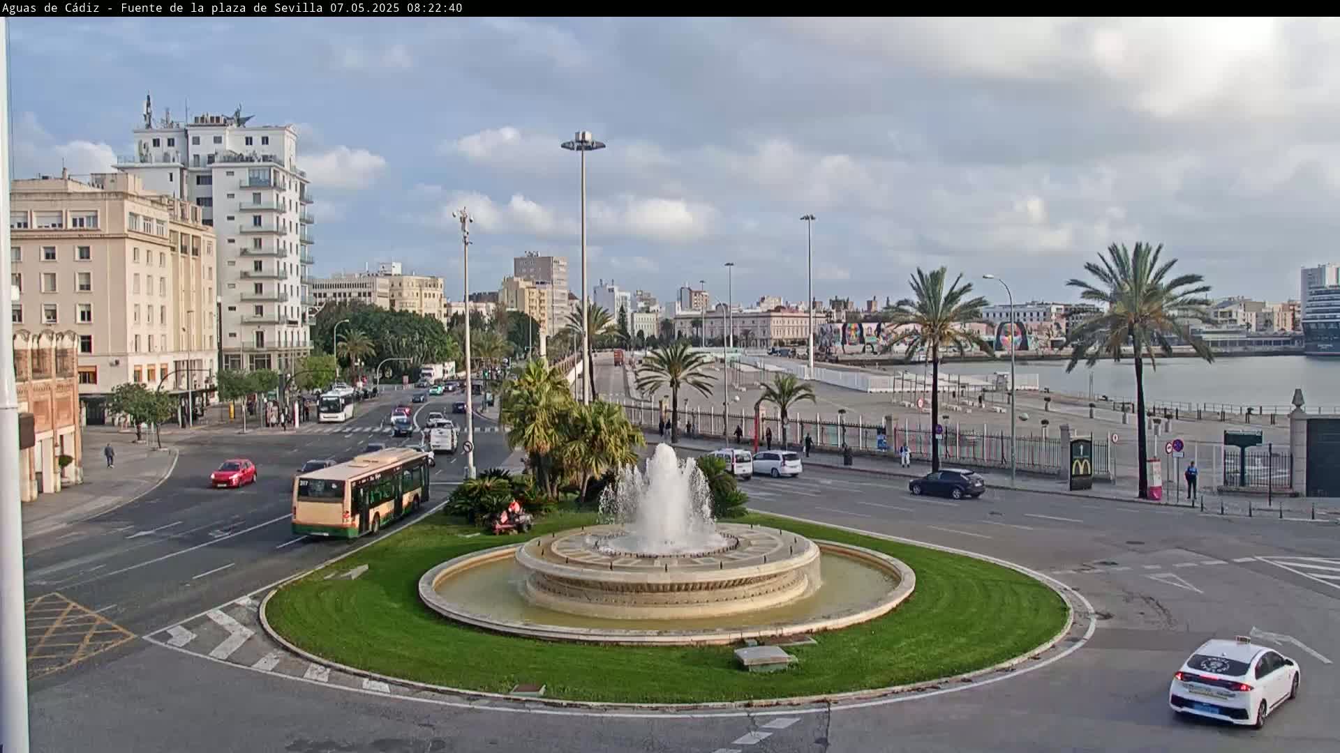 A city roundabout with a fountain in the center, surrounded by buildings, palm trees, and vehicles, under a partly cloudy sky.