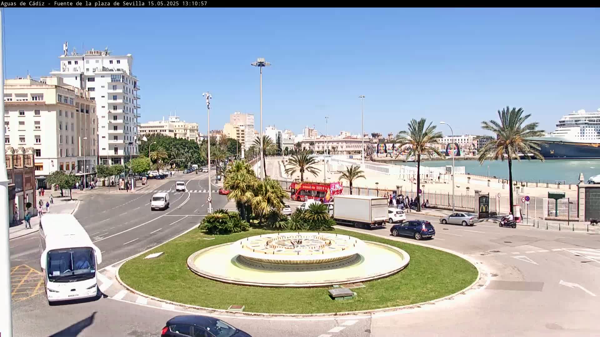 A large circular fountain sits in a traffic circle next to a waterfront area with palm trees, buildings, and a cruise ship under a clear blue sky.