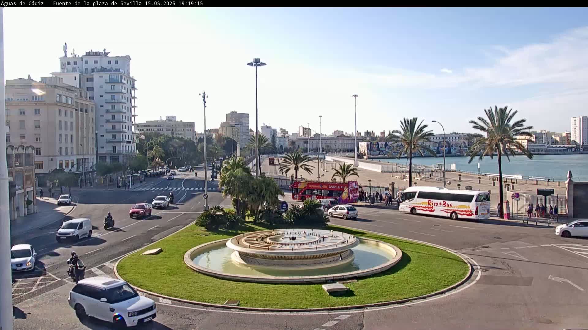 A city roundabout with a fountain in the center, surrounded by roads with cars and buses, palm trees, and buildings, all under a sunny sky.