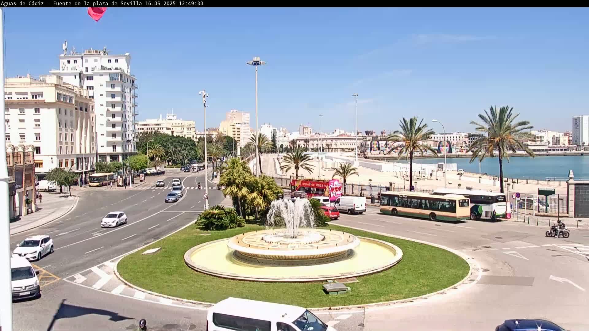 A sunny day reveals a city streetscape featuring a roundabout with a fountain, palm trees, buildings, cars, buses, and a beach in the background.