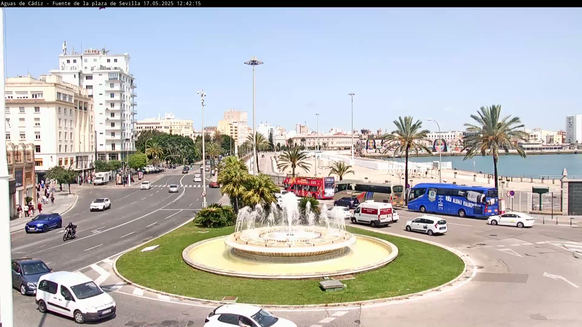 A traffic circle with a fountain in the center is surrounded by buildings, palm trees, and vehicles on a sunny day.