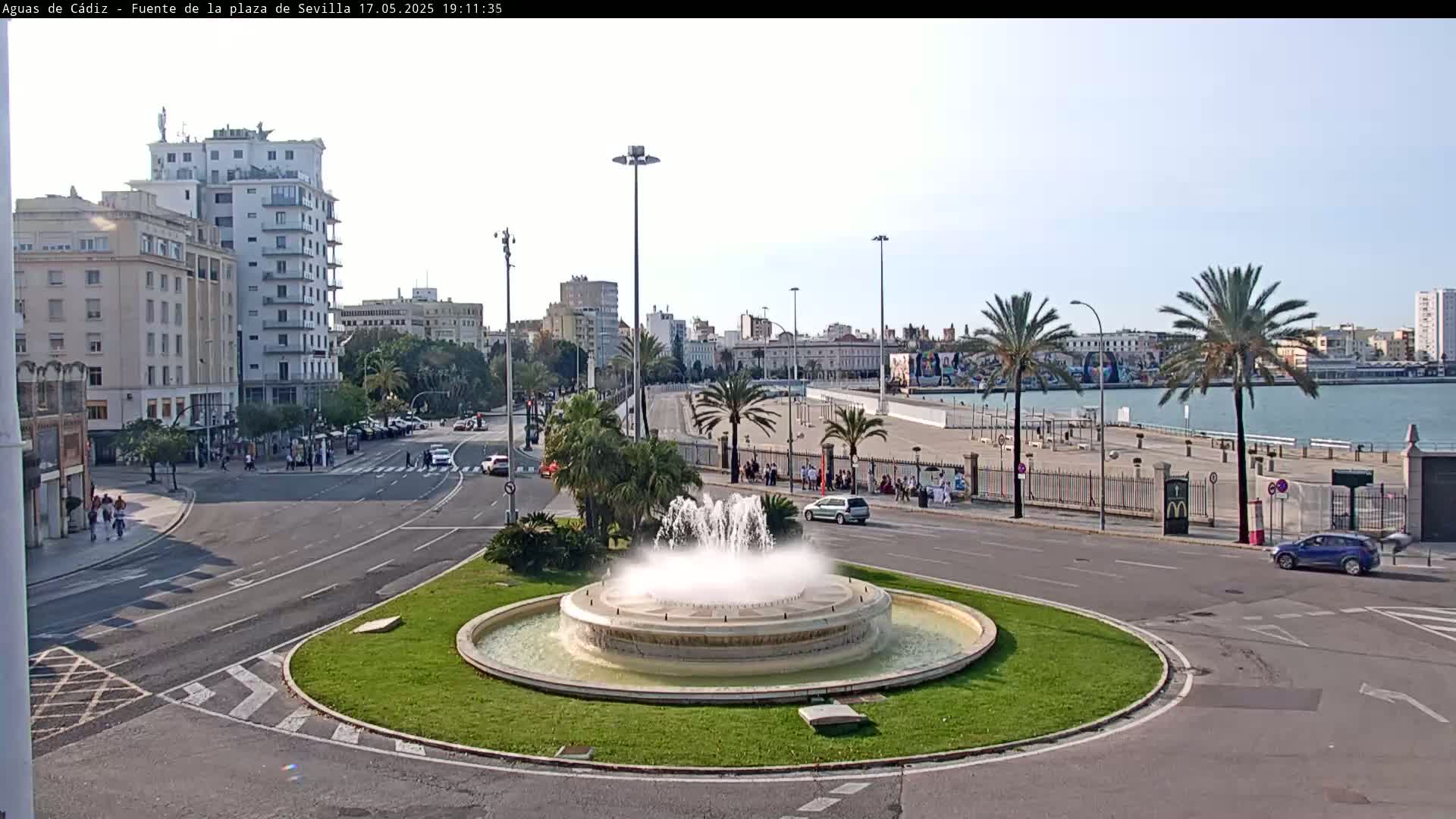 A circular fountain sprays water in a city square overlooking a harbor on a partly sunny day.