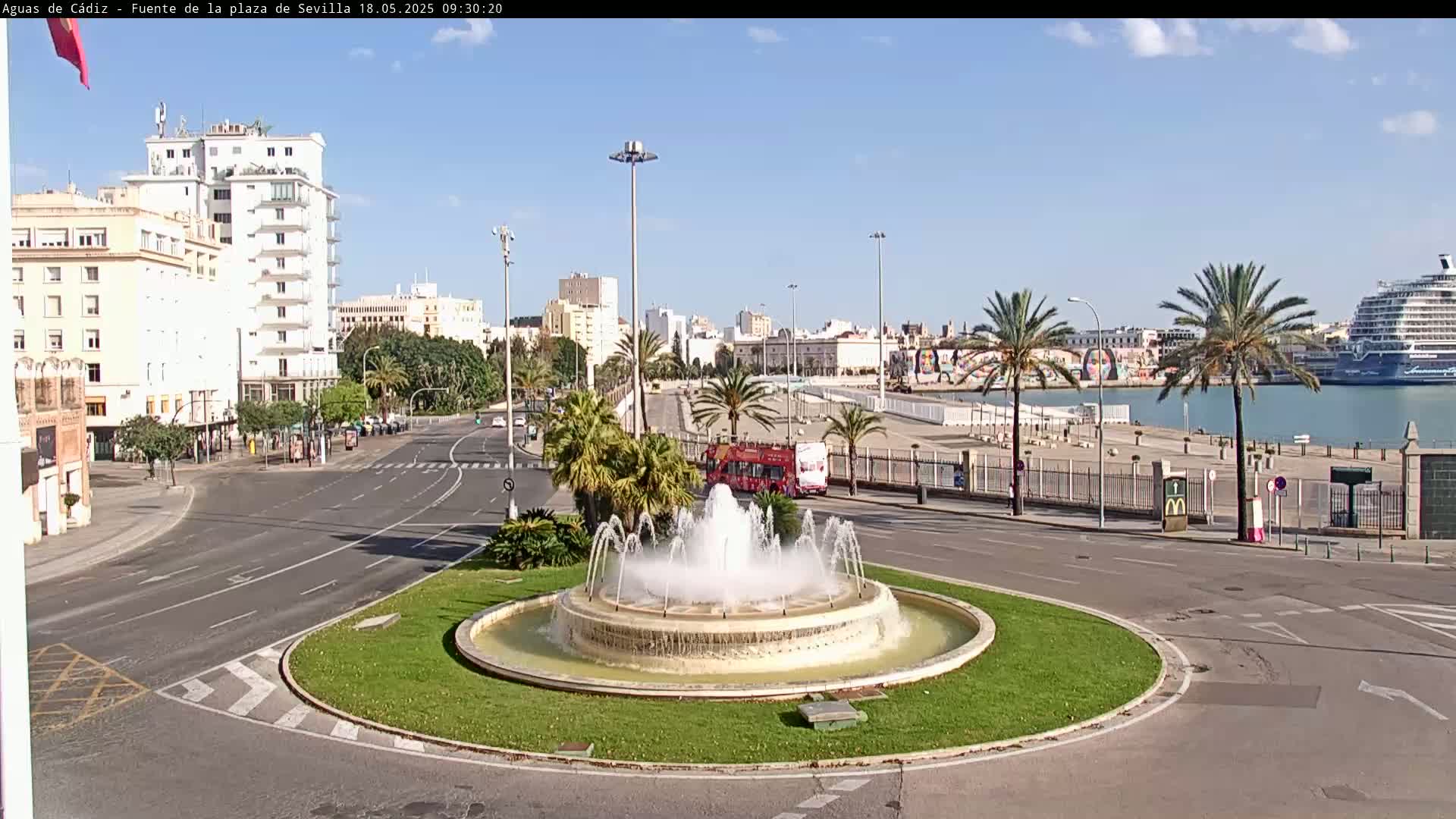 A large fountain sits in a traffic circle overlooking a harbor with a cruise ship under a clear, sunny sky.