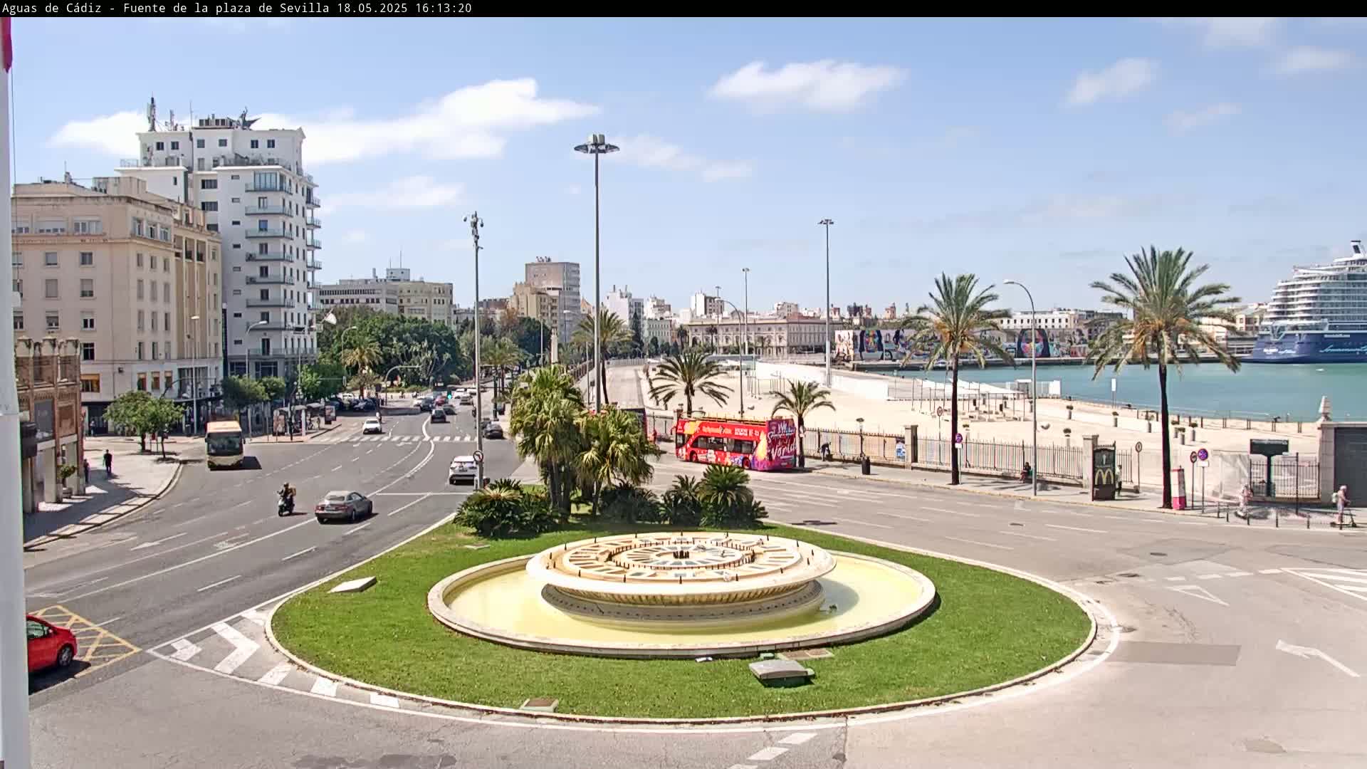 A city roundabout with a central fountain is surrounded by roads, buildings, palm trees, and a waterfront with a cruise ship under a partly cloudy sky.