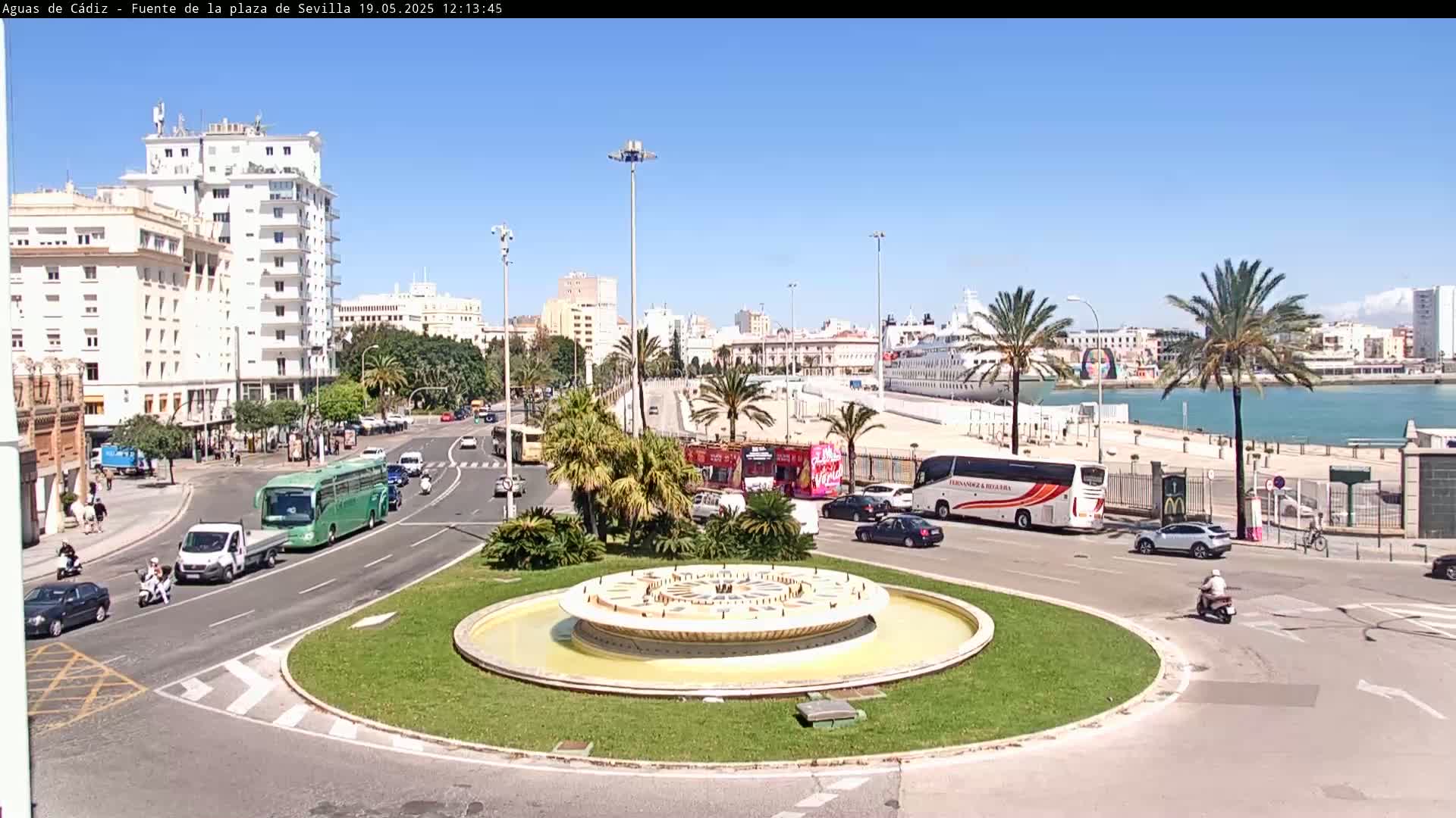 A circular fountain sits in the center of a traffic circle, surrounded by city streets, buildings, palm trees, and a cruise ship docked at a waterfront on a sunny day.