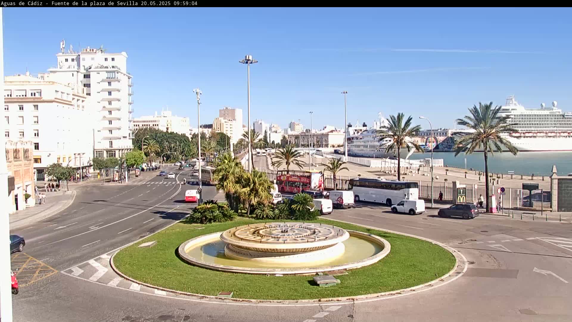 A circular fountain sits in a traffic circle near a harbor where several cruise ships are docked under a clear blue sky.