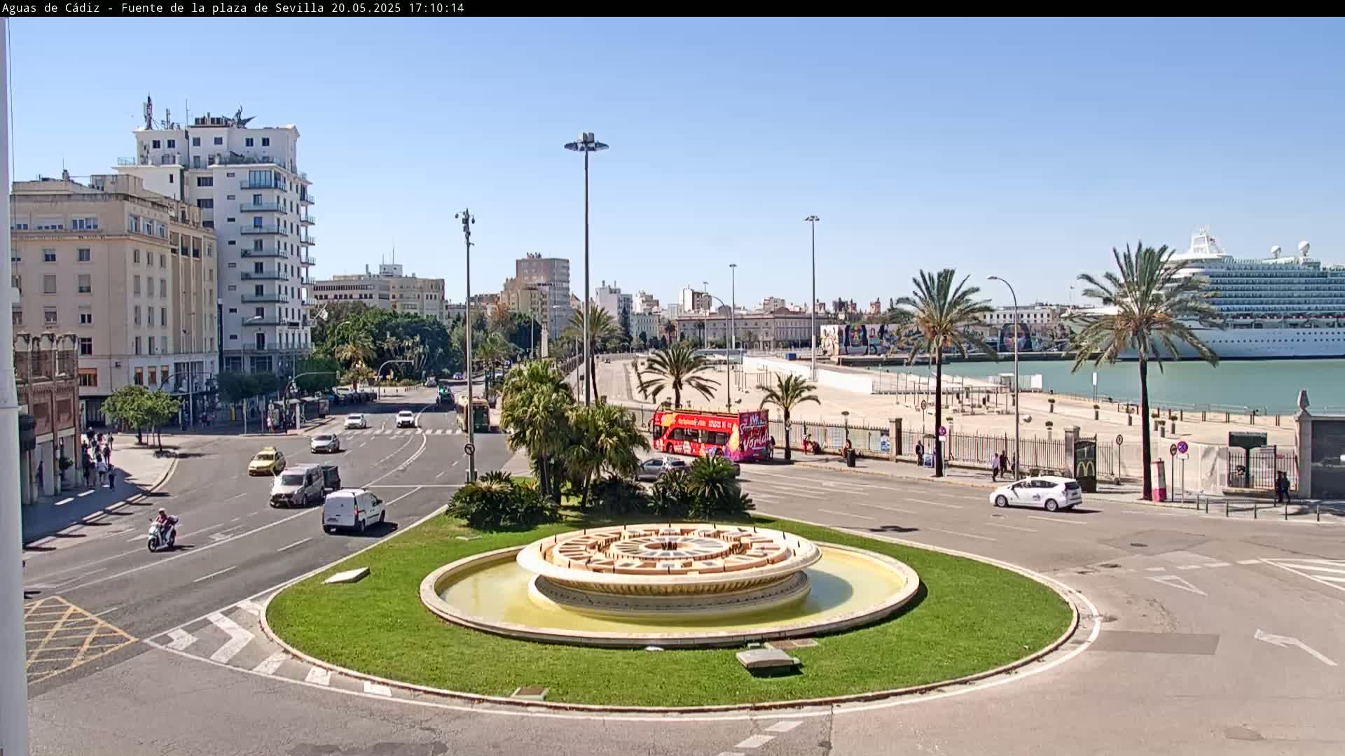 A circular fountain sits in a traffic circle, surrounded by buildings, palm trees, vehicles, and a large cruise ship docked in a harbor under a sunny sky.